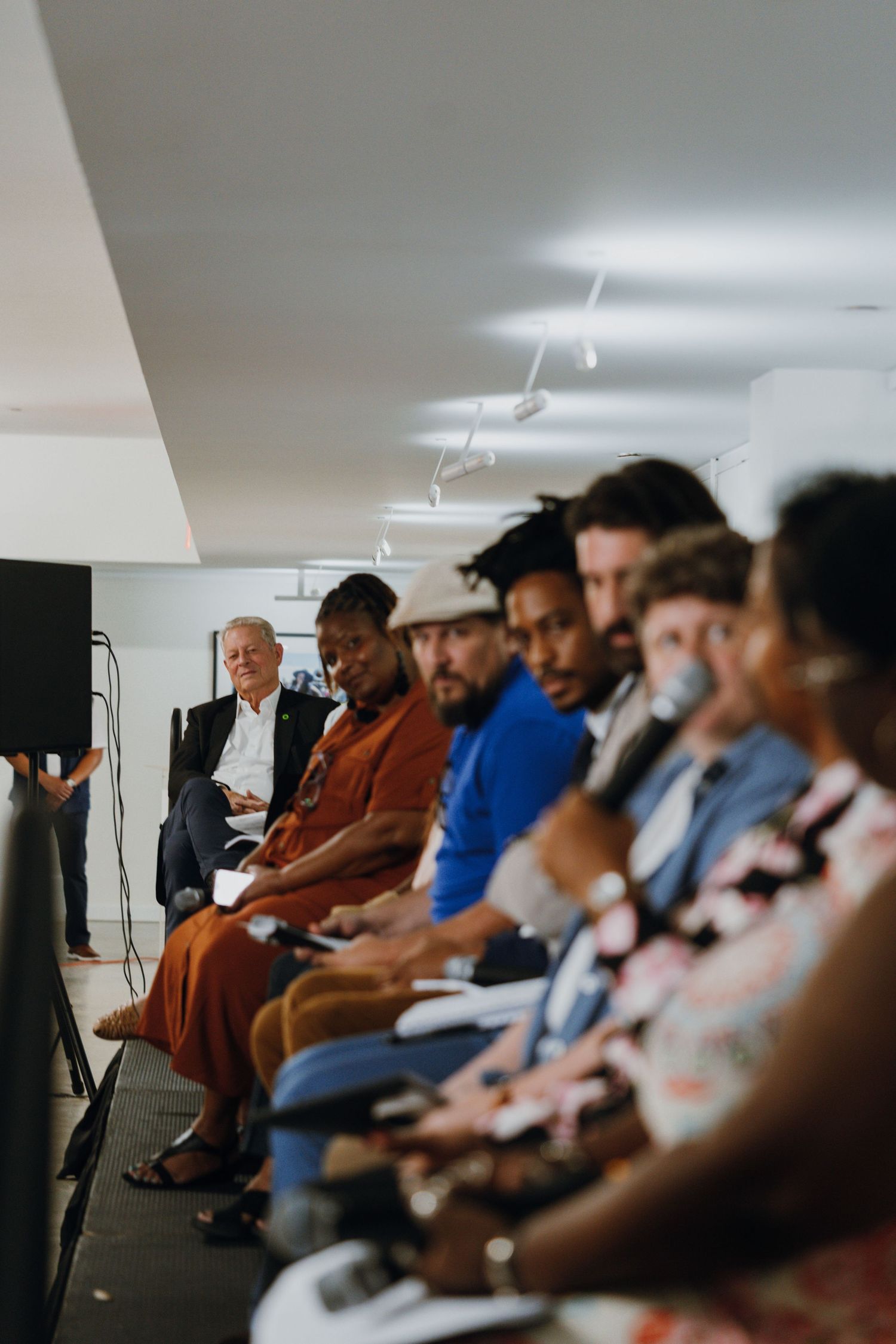 A diverse panel of speakers sits in a row on a stage during an indoor community discussion event.