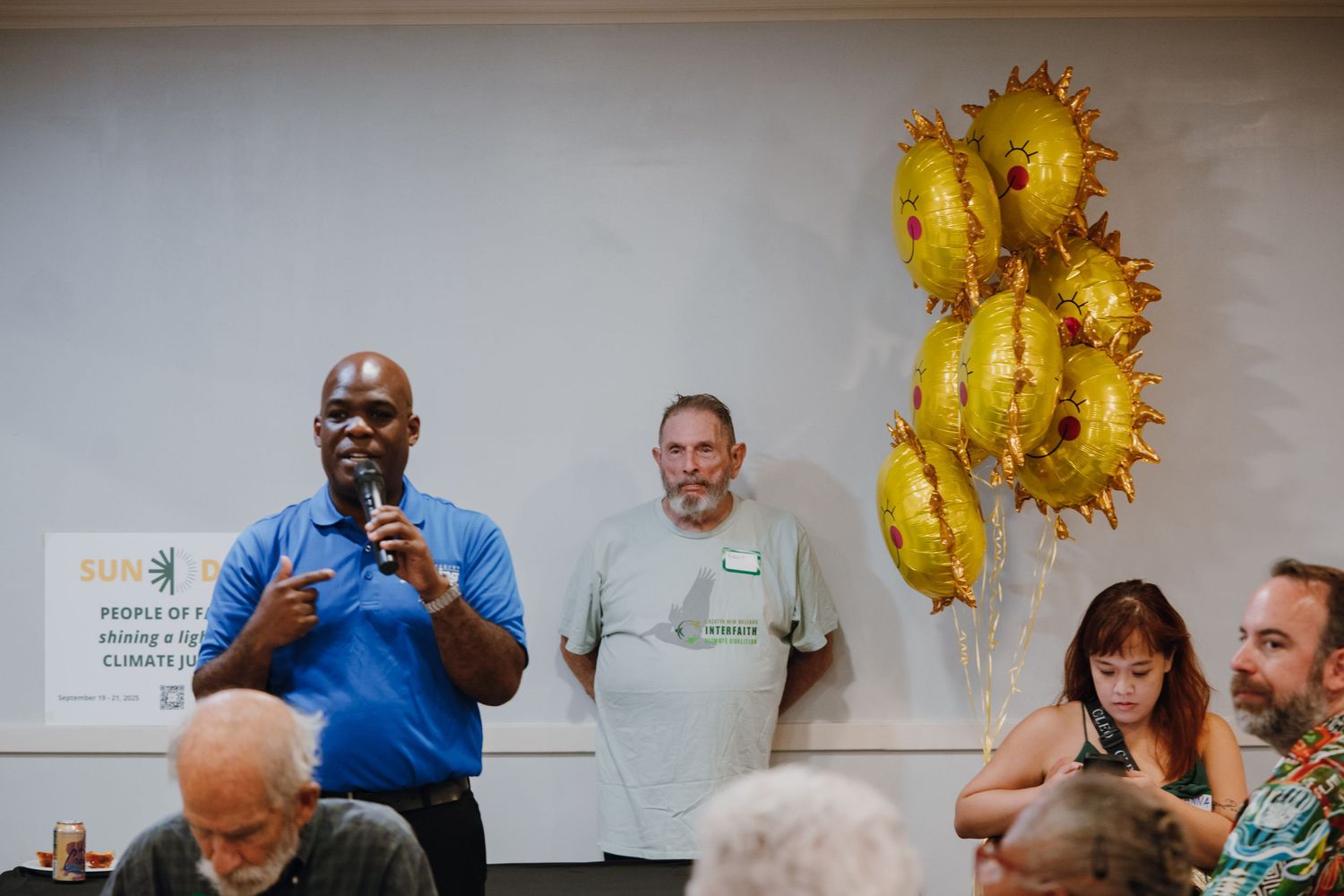 Man speaking at microphone during community meeting with yellow sun balloons and attendees seated.