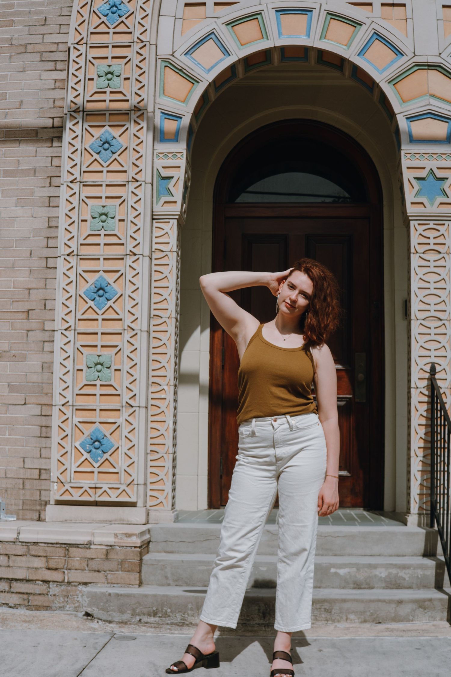 Young woman in mustard tank top and white jeans posing before ornate arched doorway with colorful tile details.