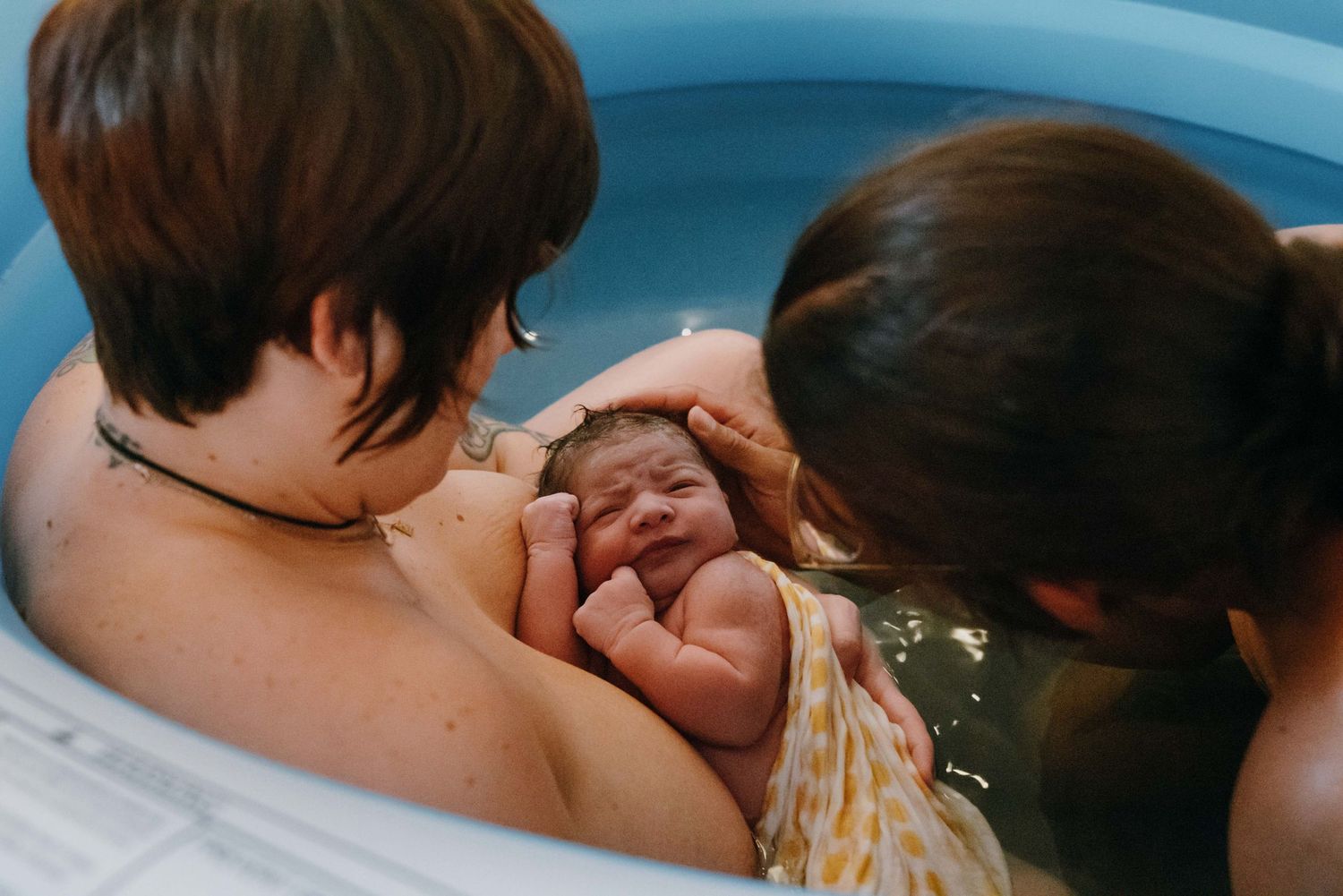 Parents cradling newborn baby together in blue inflatable water birth pool.