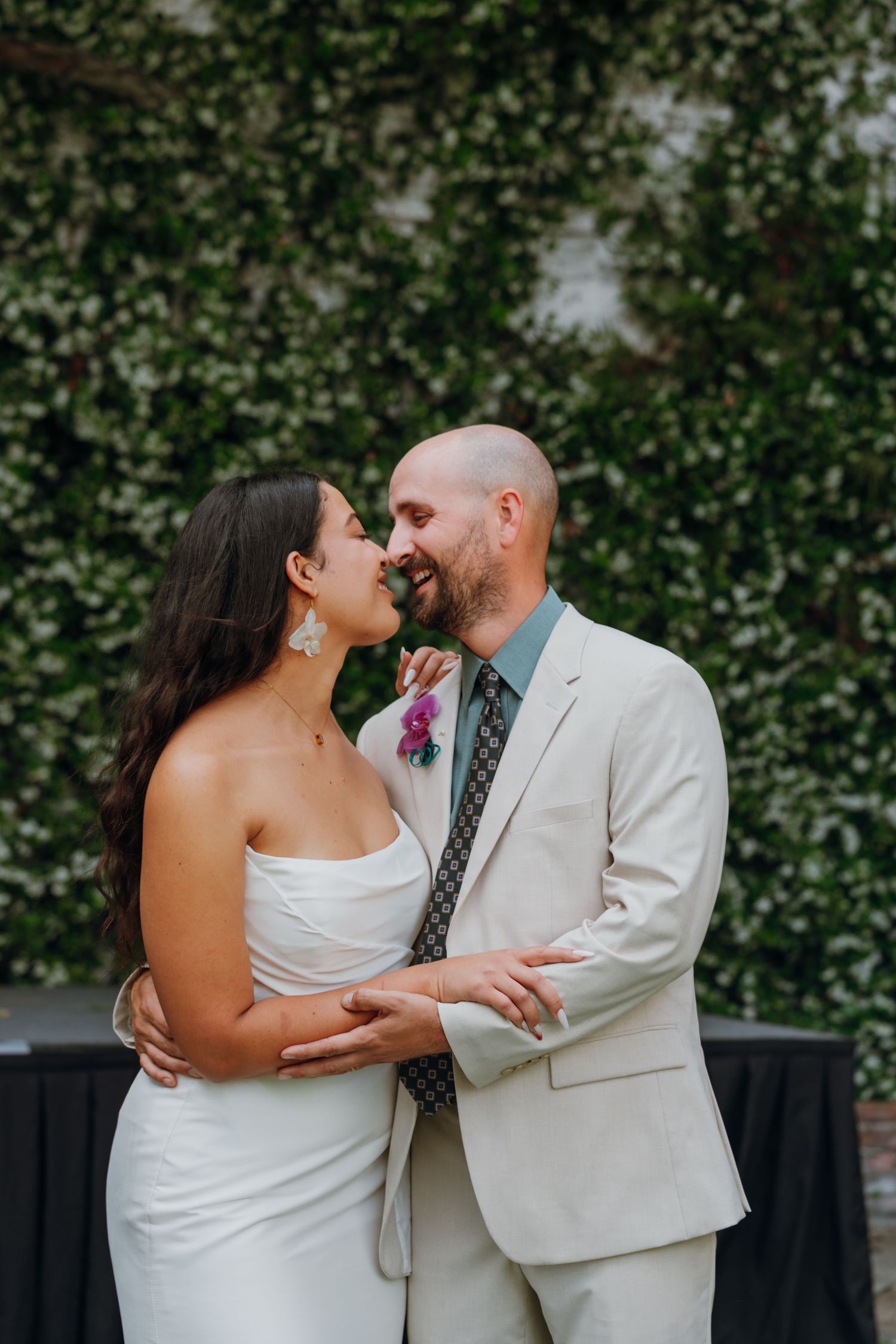 Bride and groom share romantic moment smiling at each other in front of green ivy wall.