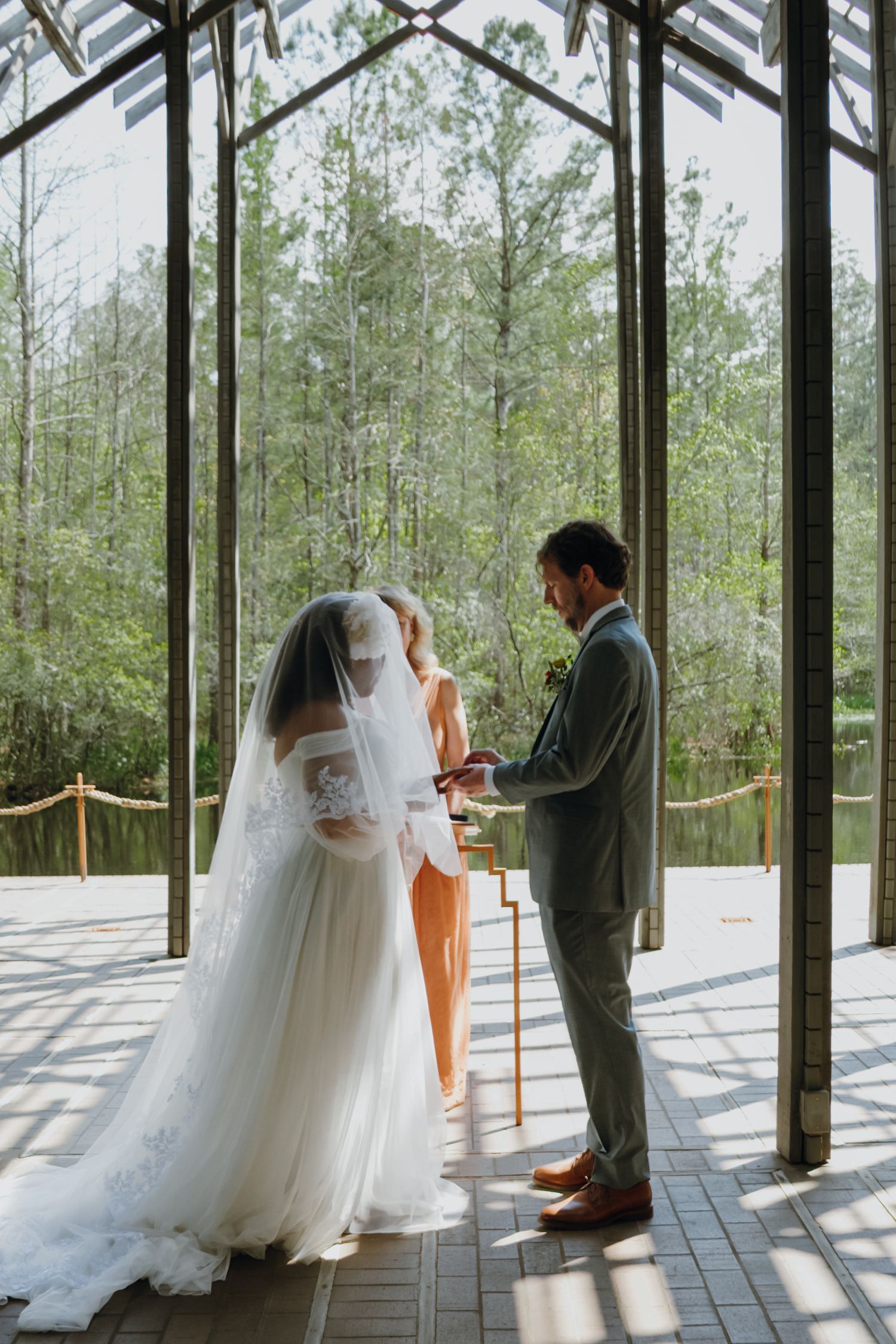 Bride and groom exchange vows at glass chapel altar with lush forest backdrop.