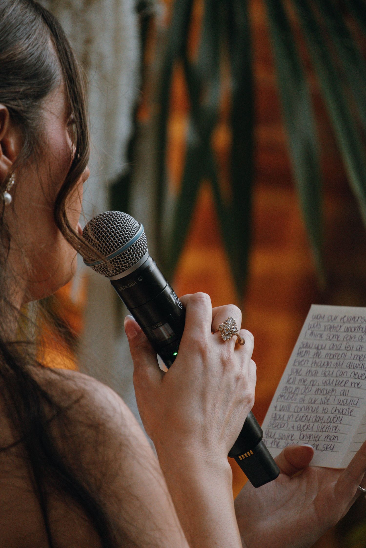 Woman reads handwritten vows into a microphone wearing a decorative ring at a wedding.