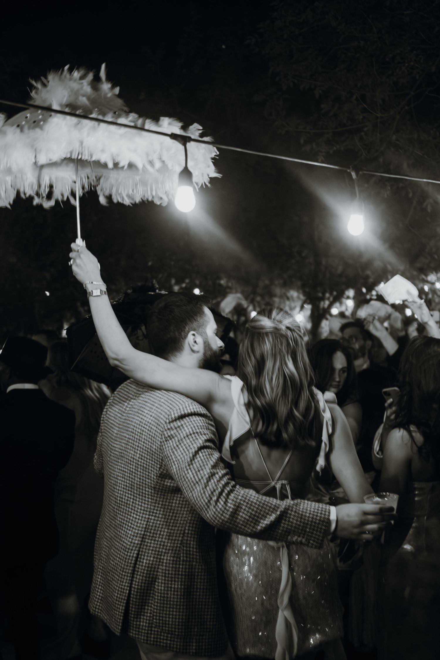 Black and white photo of bride and groom dancing with feather umbrella at outdoor wedding reception.