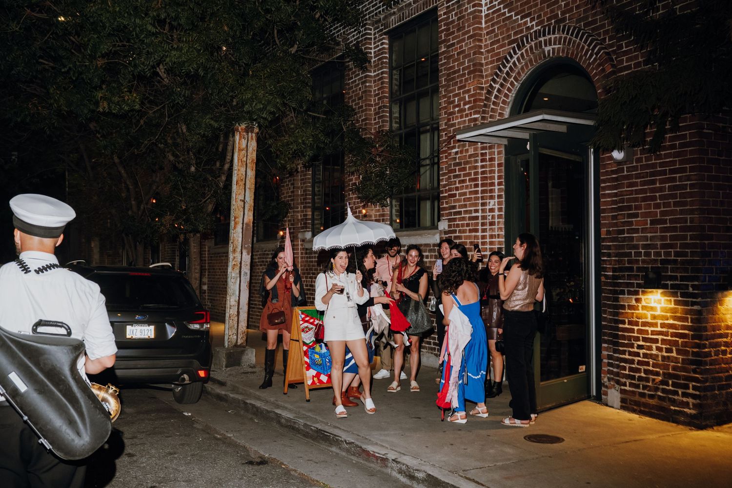 A group of people gather outside a brick building at night while a security guard stands nearby.