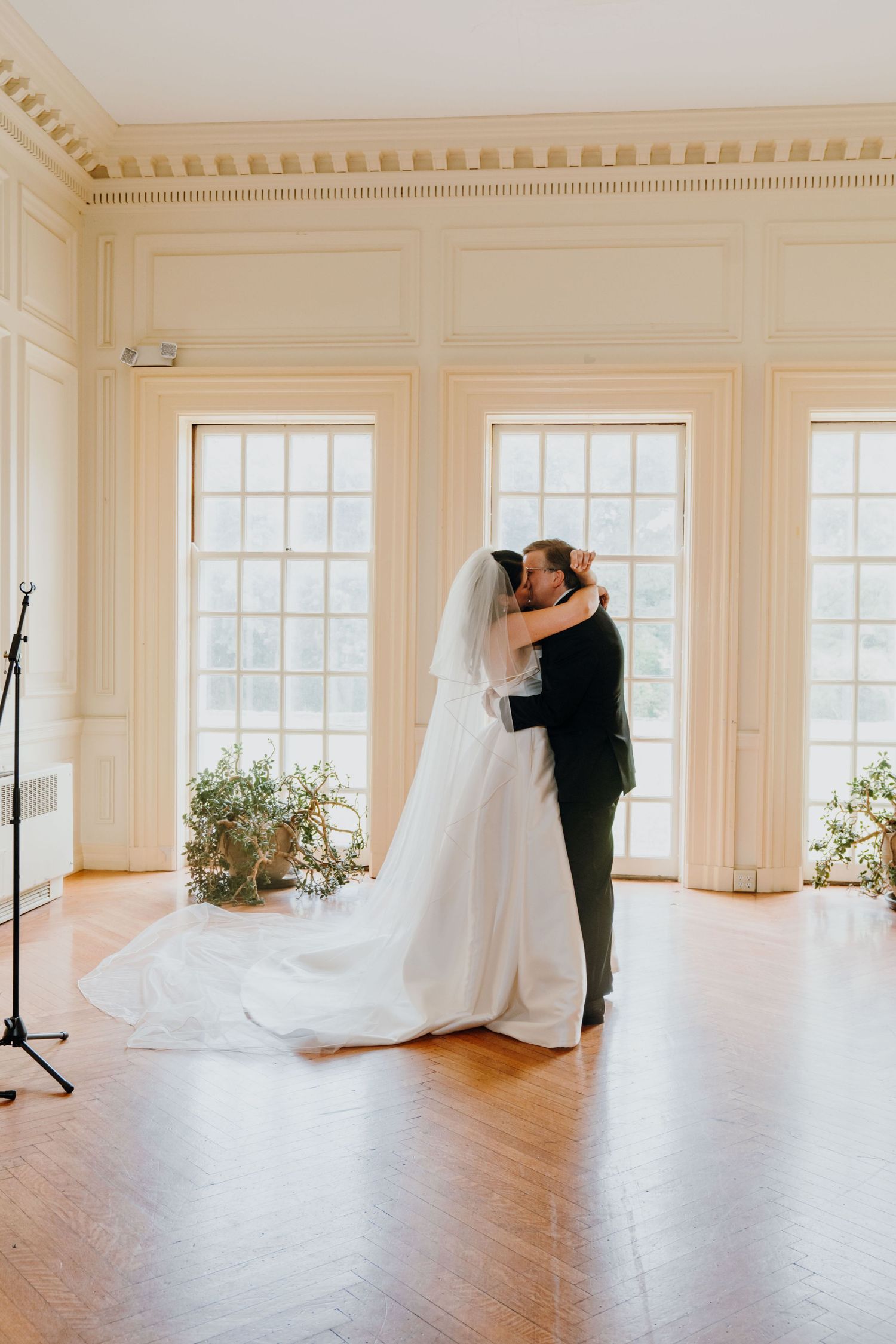 Bride and groom share a tender first dance embrace in an elegant ballroom with tall windows and warm natural light.