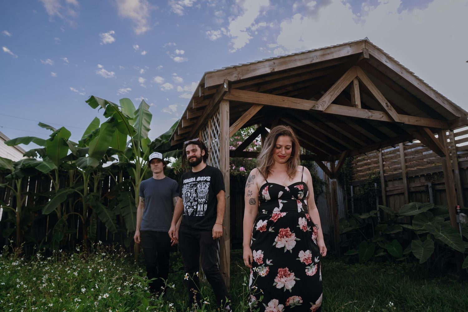 Three musicians pose outdoors near a wooden pergola surrounded by tropical plants under a blue sky.