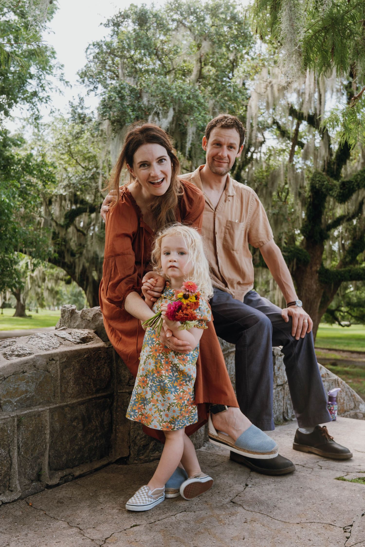 Family portrait of parents and toddler girl holding flowers, sitting outdoors among moss-draped oak trees.