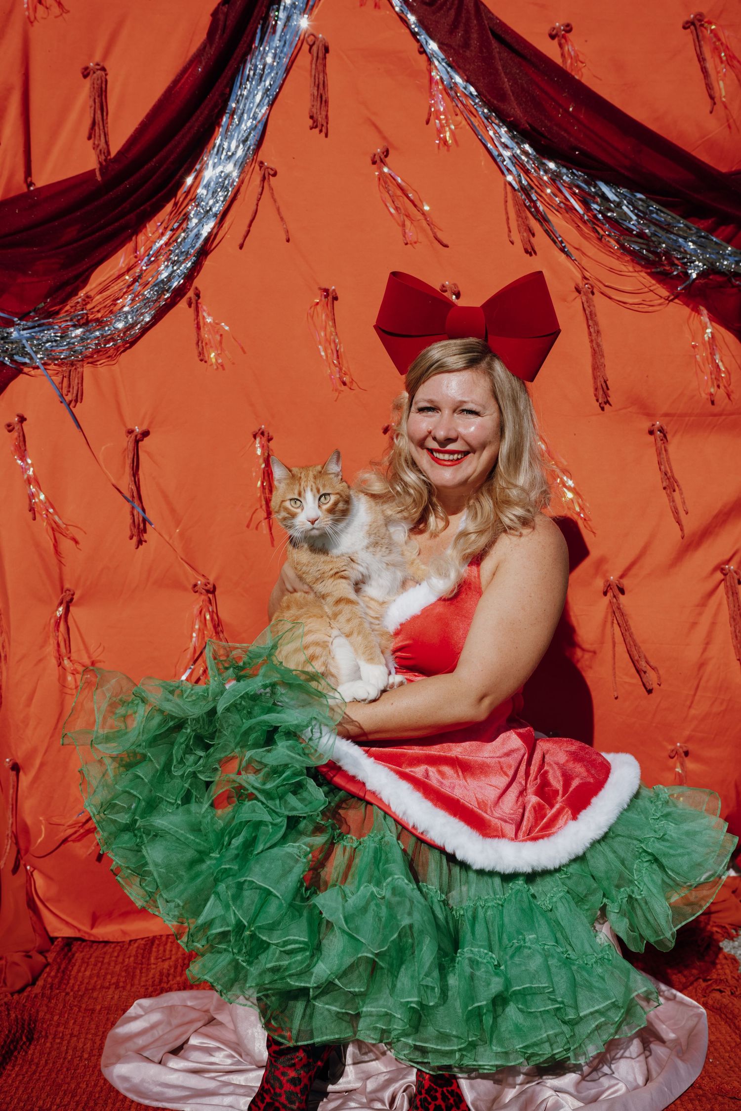 Woman in red Santa dress with green tulle skirt holds orange tabby cat, posing before festive red backdrop.
