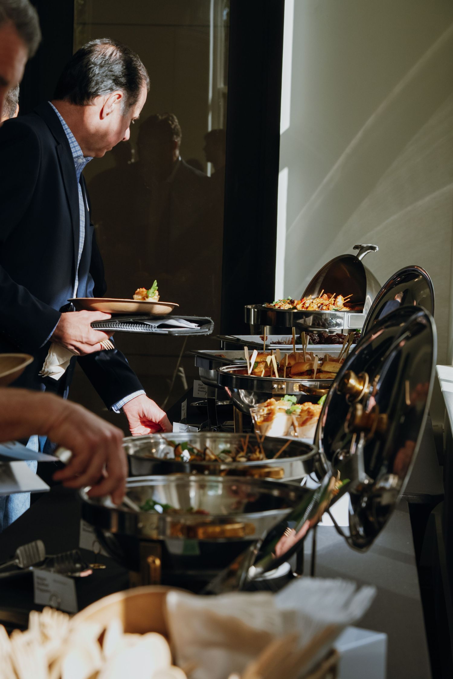 Man in suit serving himself at an elegant buffet with silver chafing dishes filled with various foods.