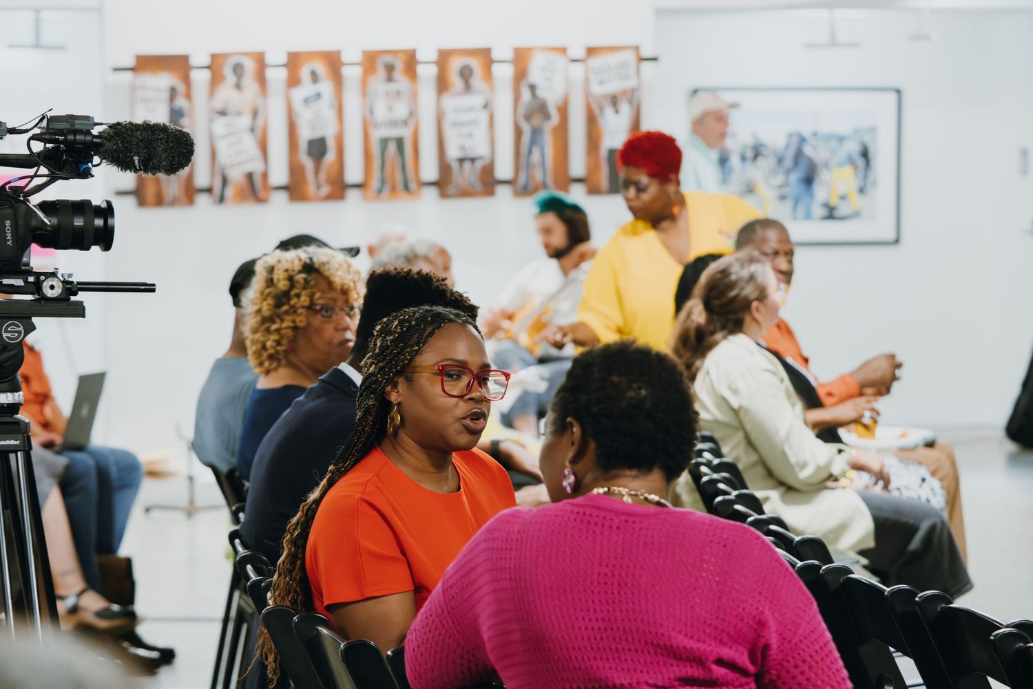 Diverse group of people seated at an indoor community event with artwork displayed on walls.