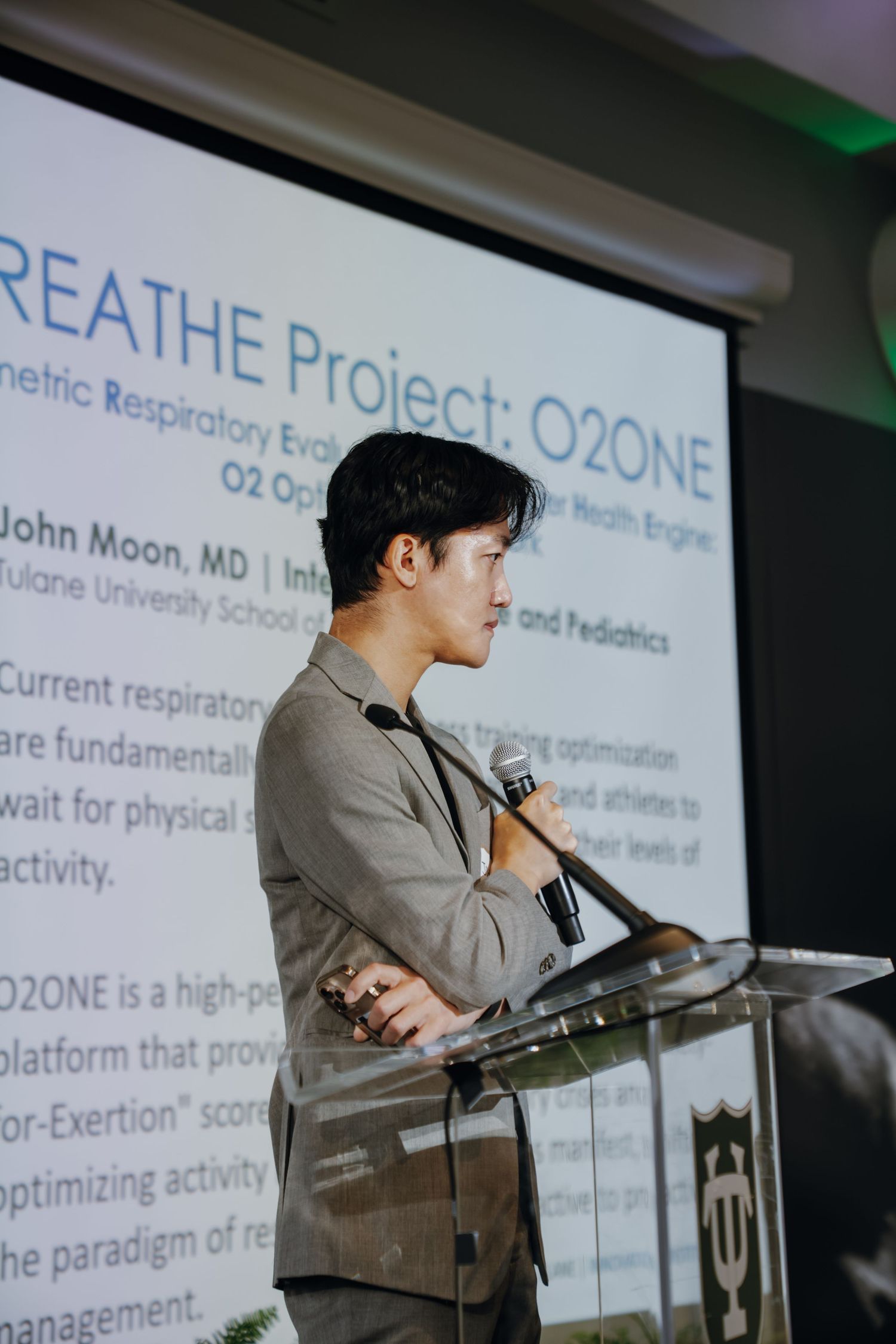 Man in gray blazer presents at podium with BREATHE Project O2ONE respiratory health slide projected behind him.
