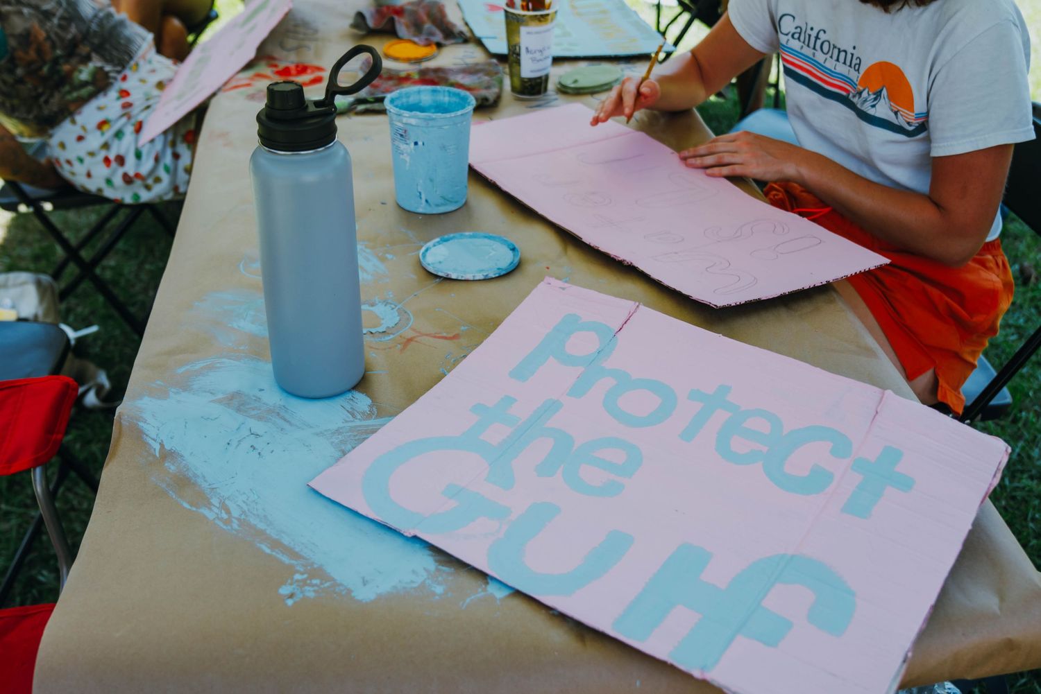 Protect the Gulf protest sign being painted in teal letters on pink poster board at a table.