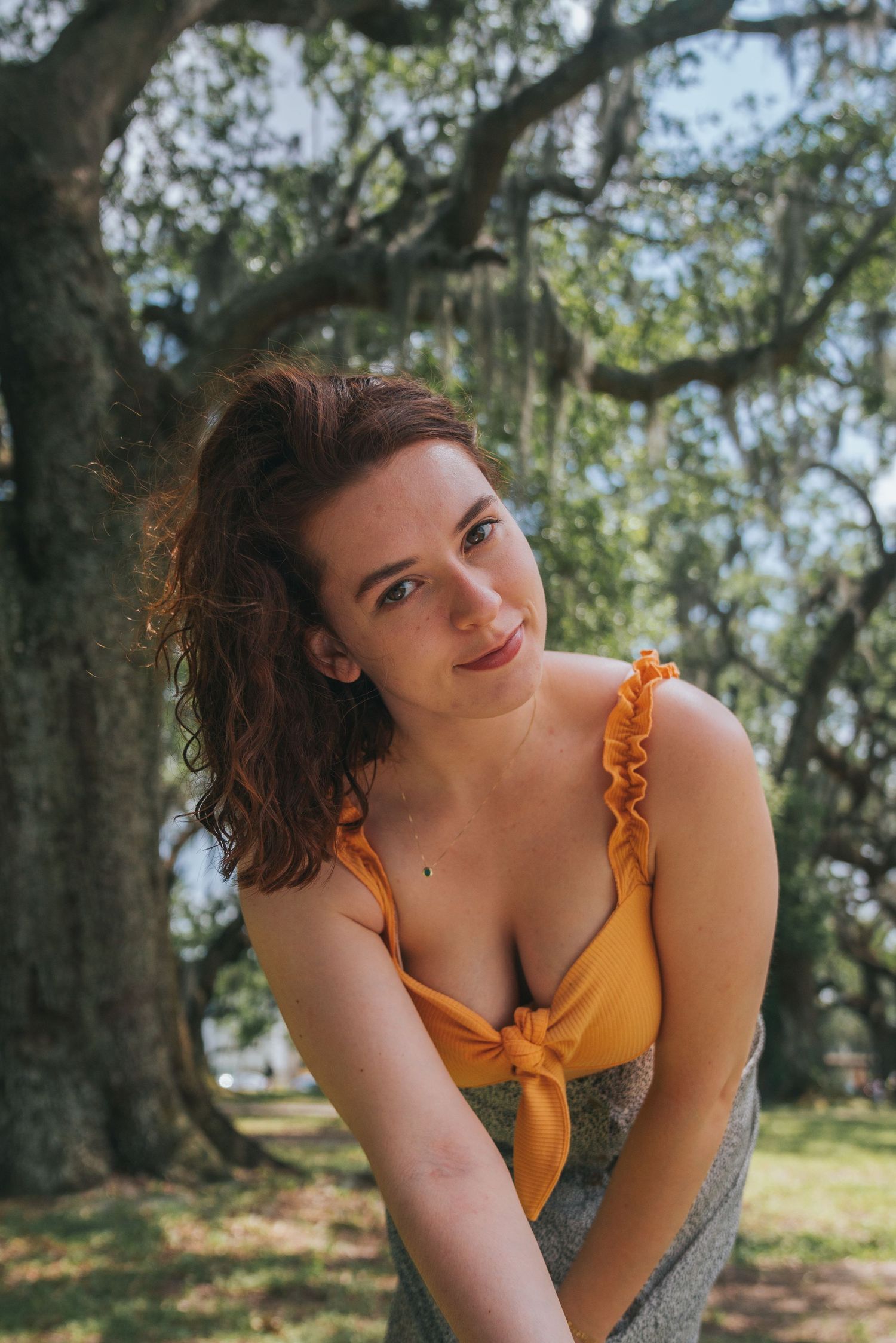 Young woman in orange tie-front top posing thoughtfully under large moss-draped trees in a park.