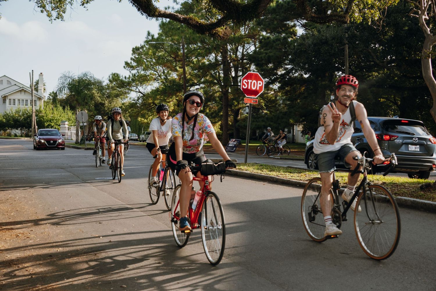 Group of cyclists riding through a tree-lined street past a stop sign on a sunny day.
