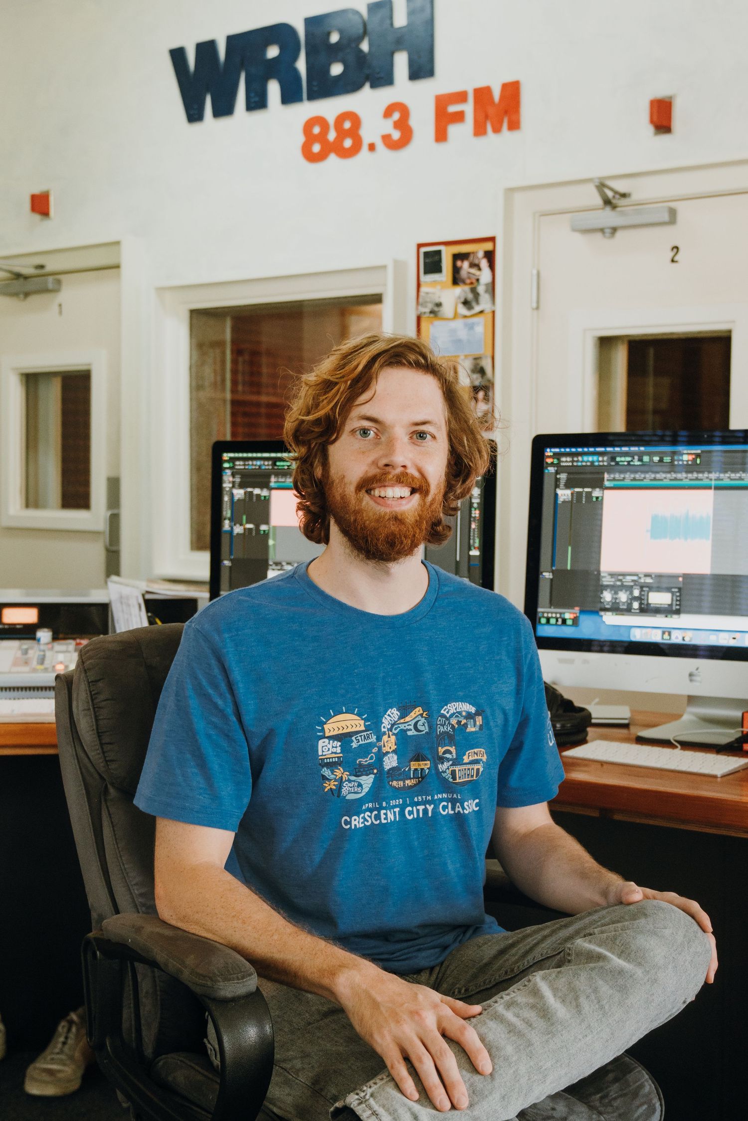 Smiling bearded man in blue t-shirt seated in front of computers at WRBH 88.3 FM radio station.