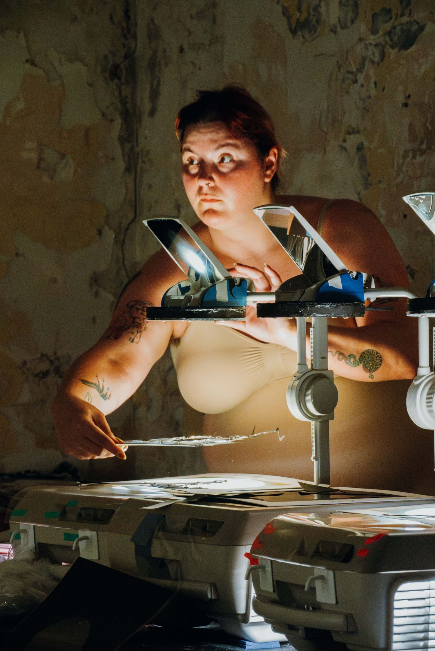 Tattooed woman operates overhead projector under dramatic lighting in worn interior.