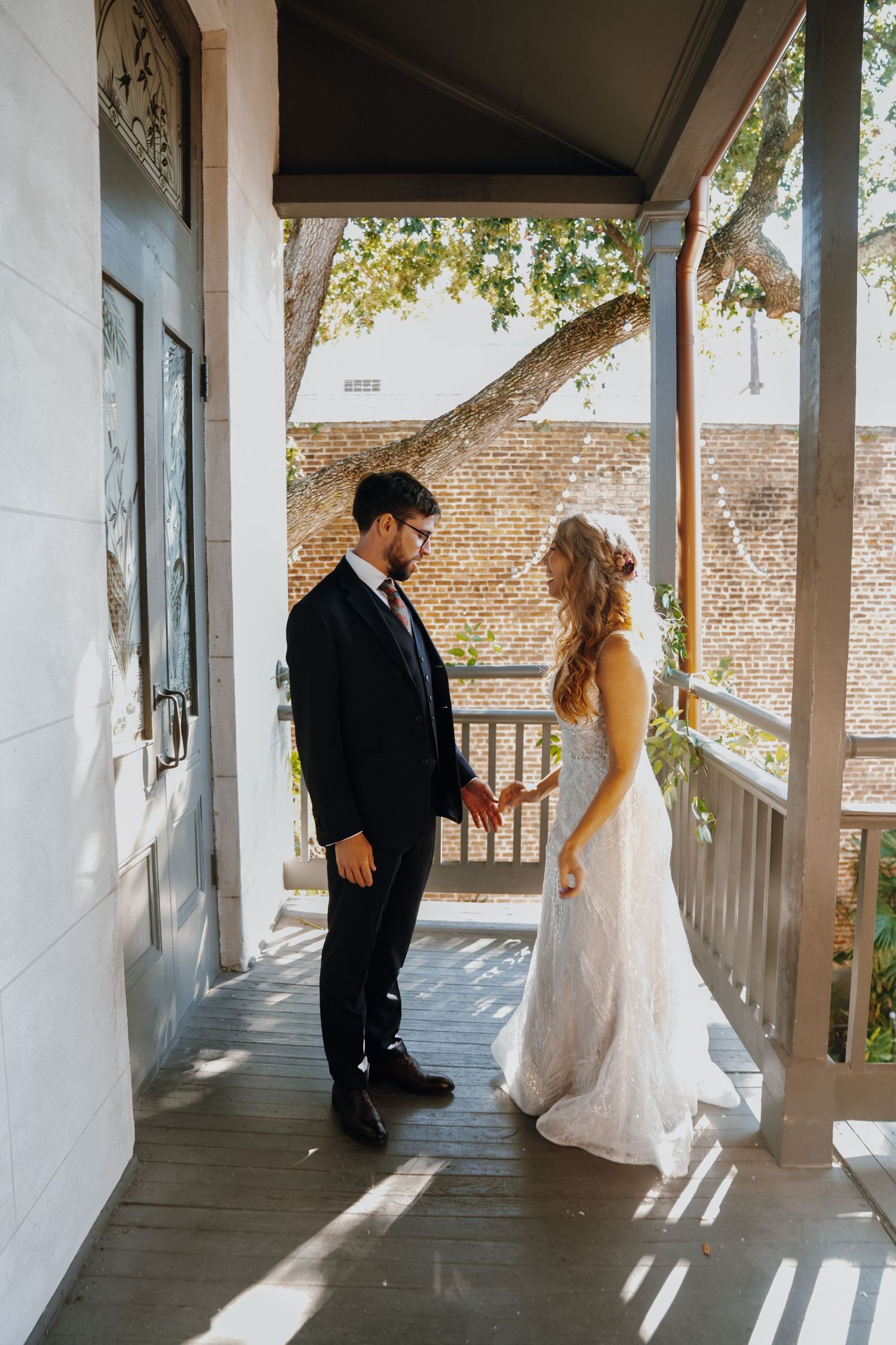 Bride in lace gown and groom in navy suit sharing first look on sunlit porch.