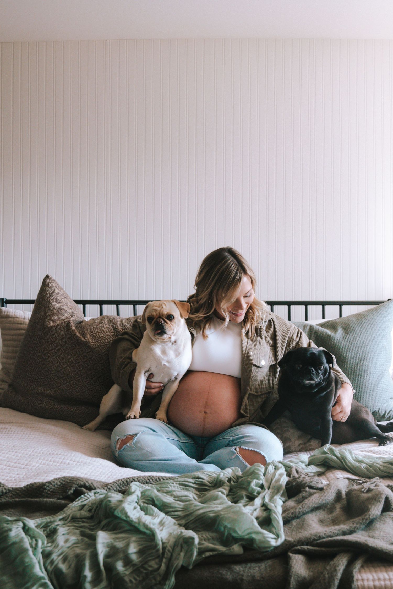 Smiling pregnant woman sitting on bed with two dogs, showing baby bump.