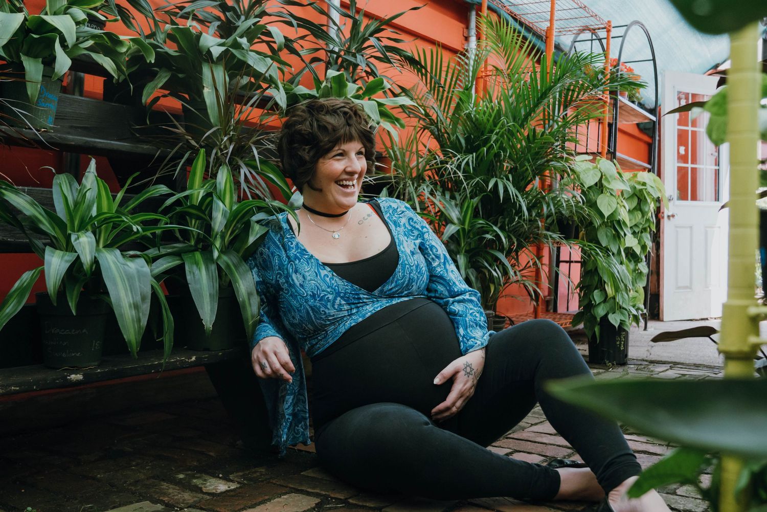 Smiling pregnant woman in blue cardigan sitting among lush green plants during maternity session. New Orleans, Louisiana, Mississippi
