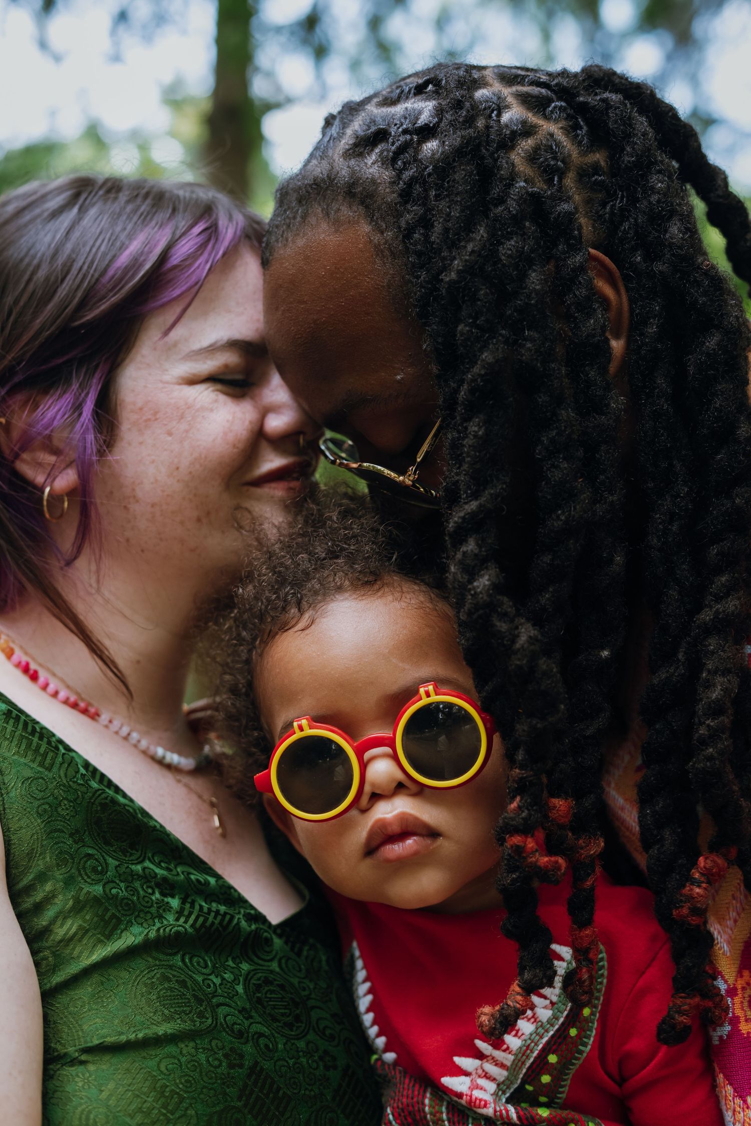 Two mothers tenderly kiss their toddler wearing yellow round sunglasses in an outdoor family portrait.