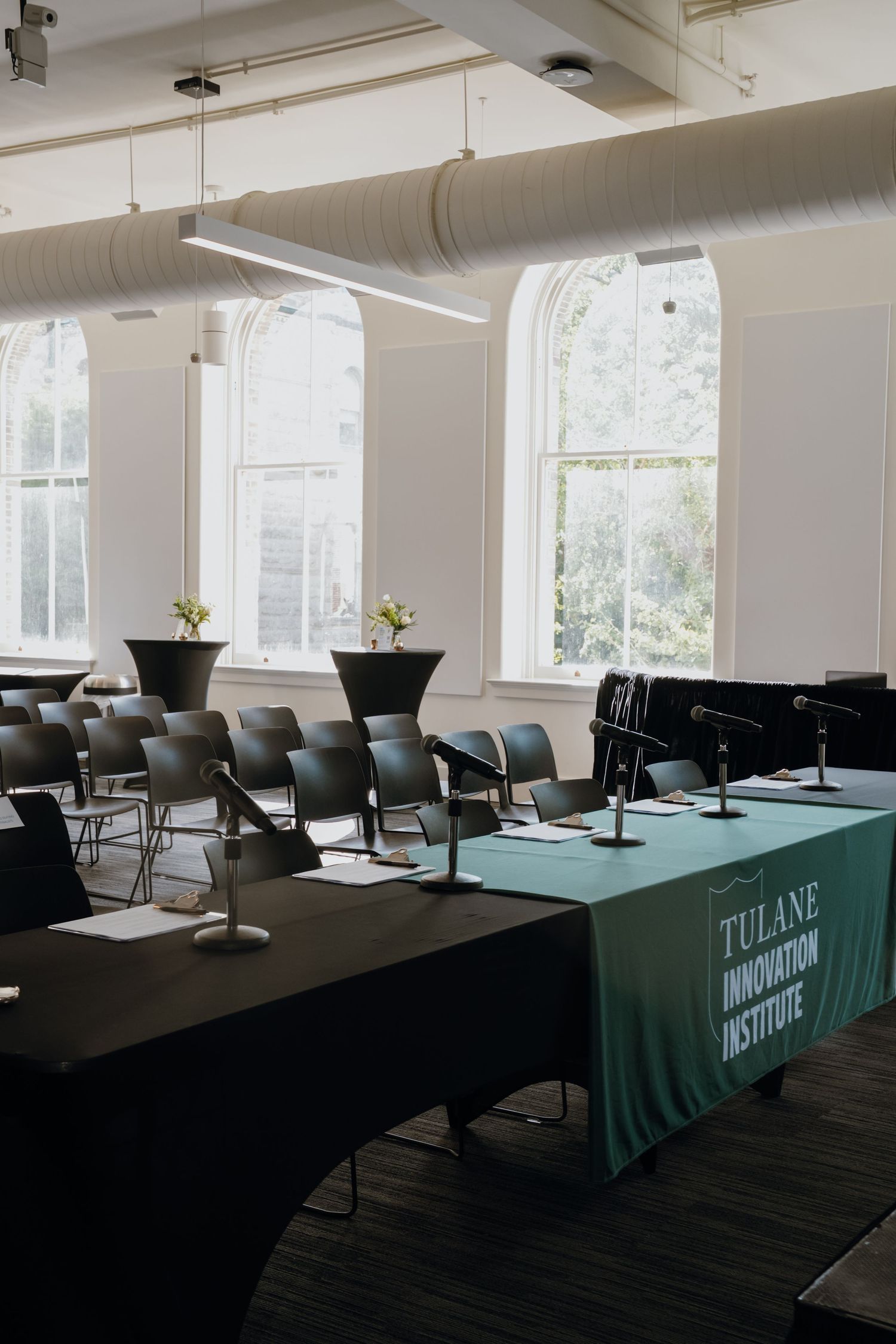 Tulane Innovation Institute conference room with black chairs, microphones, and green branded tablecloth near arched windows.