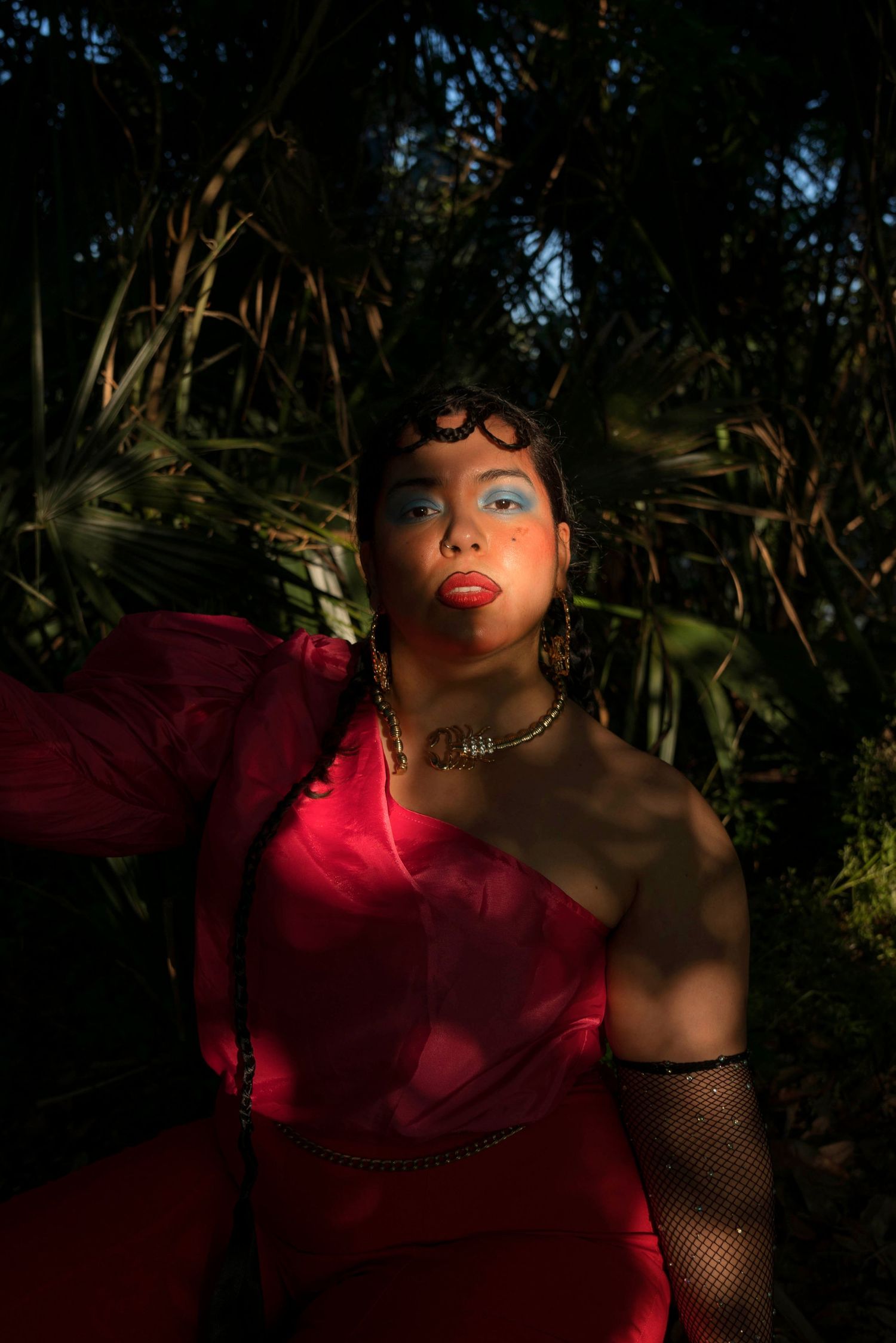 Woman in red asymmetric dress with fishnet gloves posing in dappled sunlight among tropical plants.
