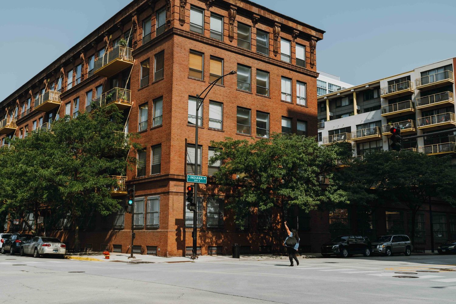 Historic red brick corner building on a sunny urban street with trees and modern buildings nearby.
