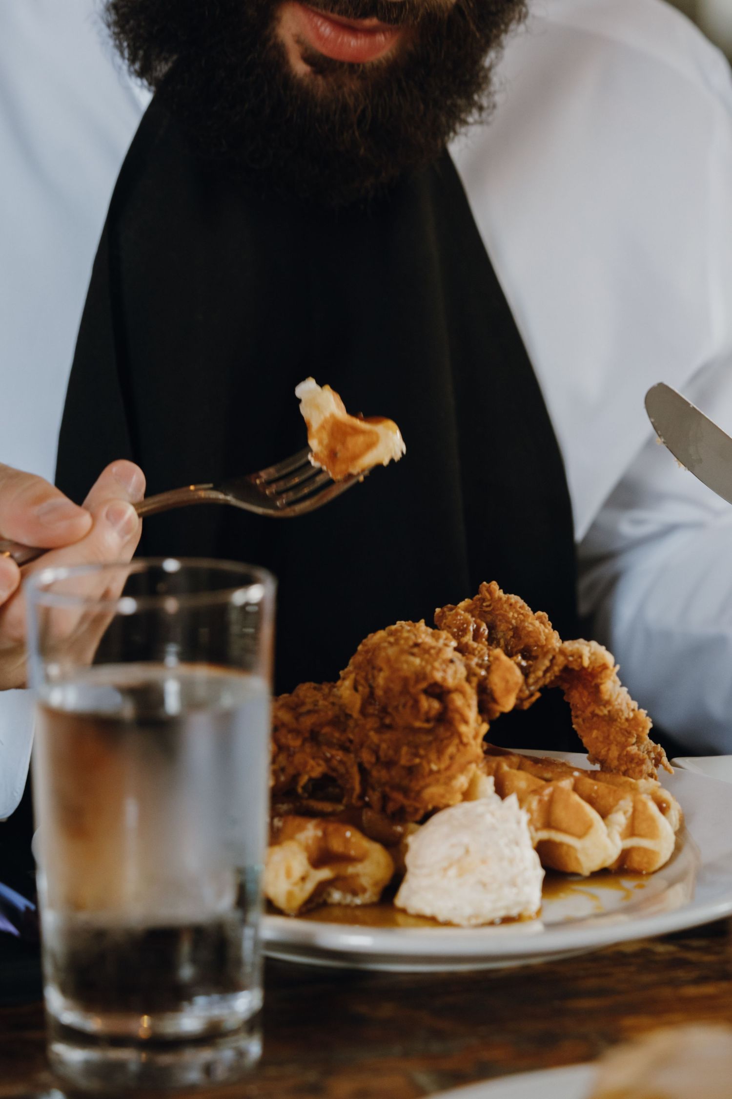 Bearded man eating crispy fried chicken and waffles with a fork at a restaurant table with a glass of water.
