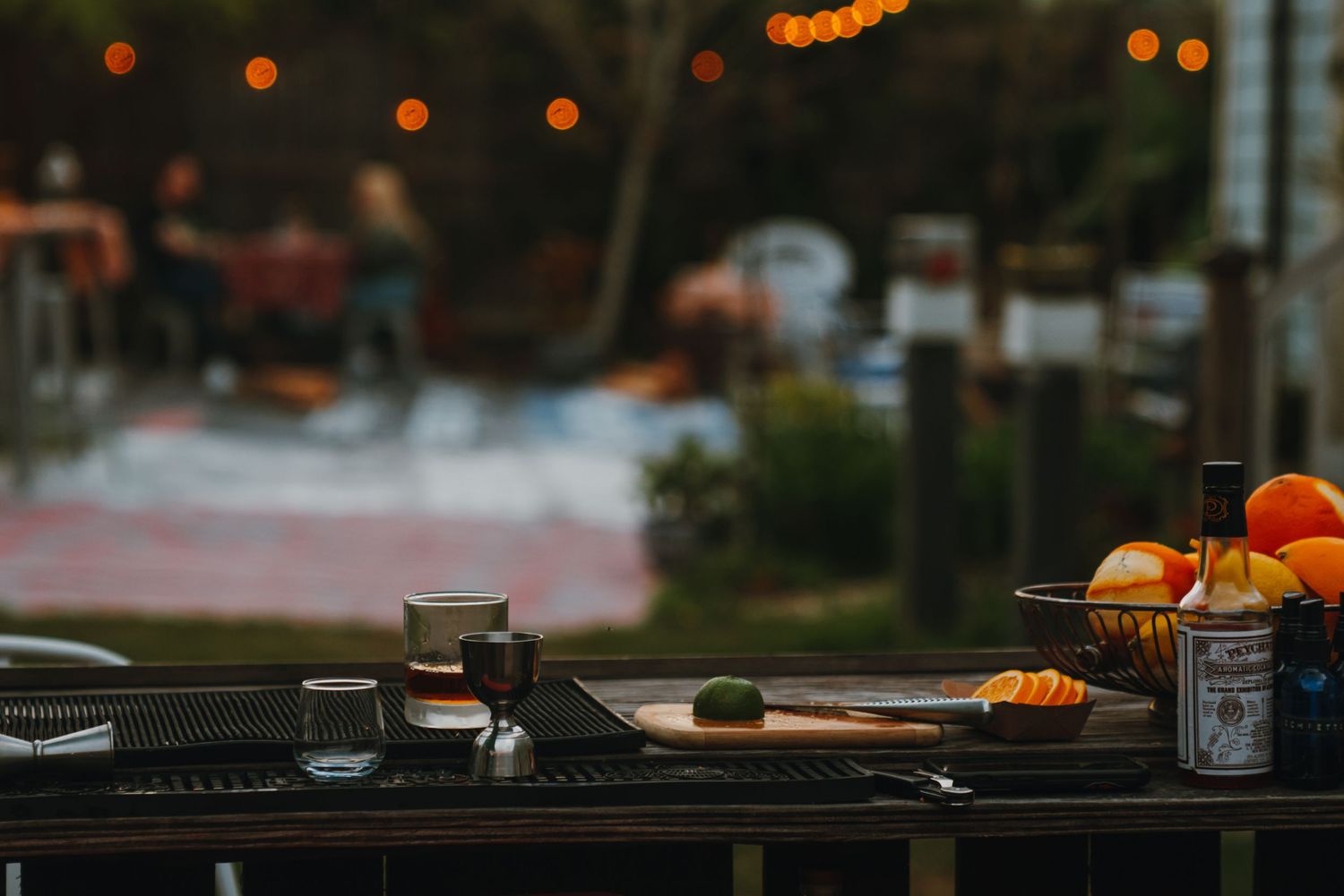Outdoor bar setup at dusk with oranges, bottles, and glowing string lights in the background.