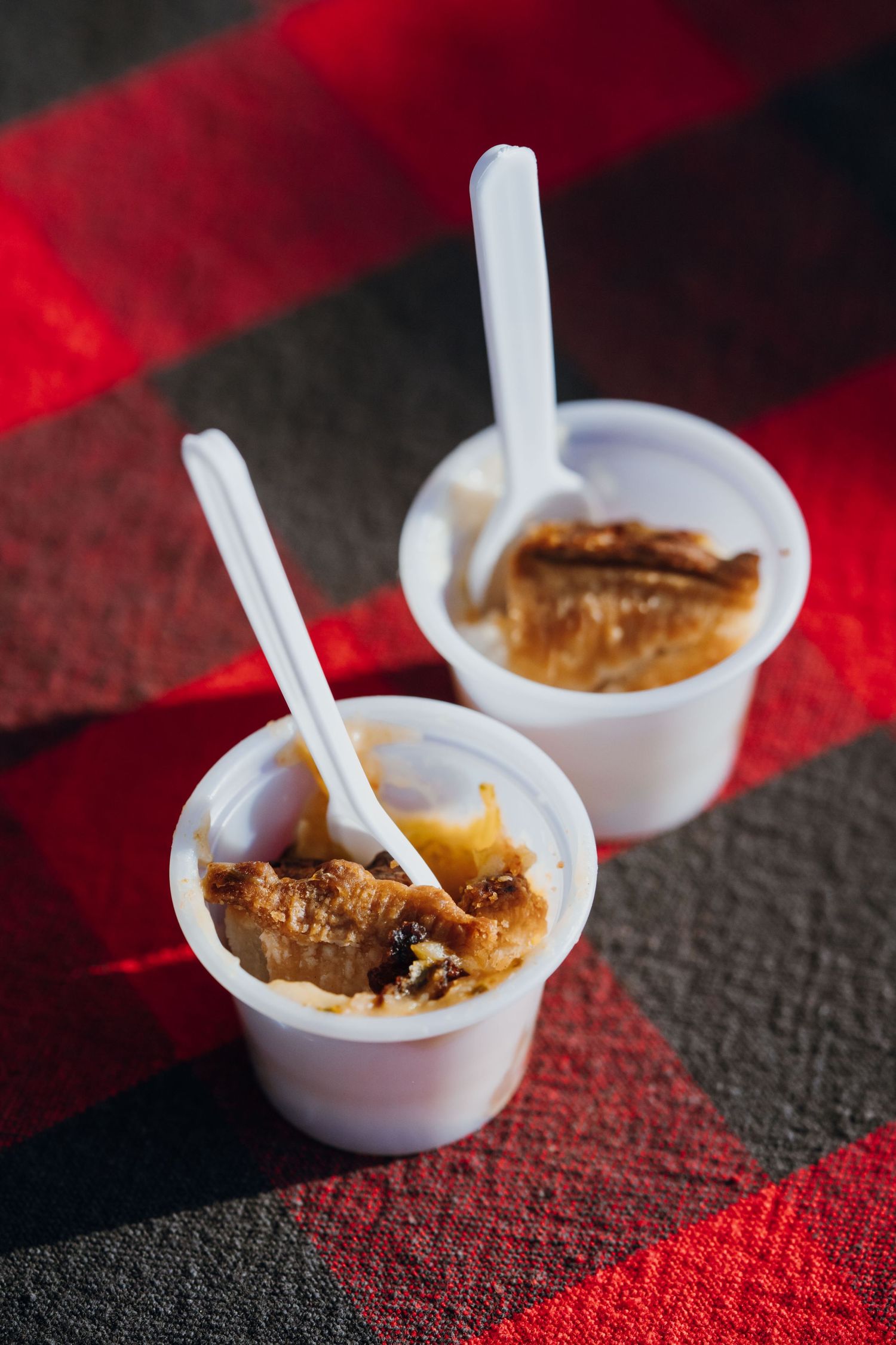 Two small white plastic cups filled with bread pudding dessert and plastic spoons on a plaid cloth.