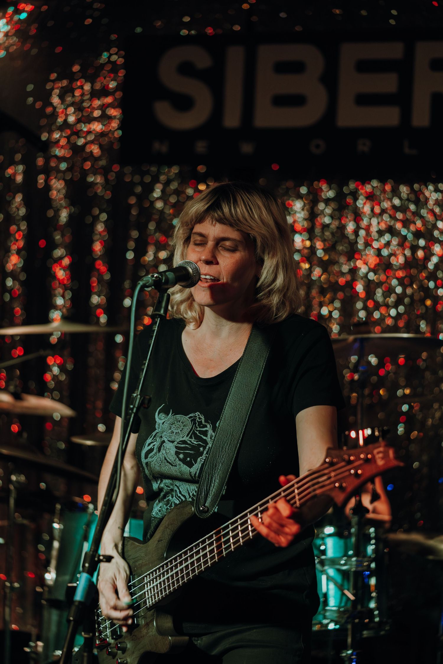 Female bassist singing into microphone on stage at Siberia venue with bokeh lights backdrop.