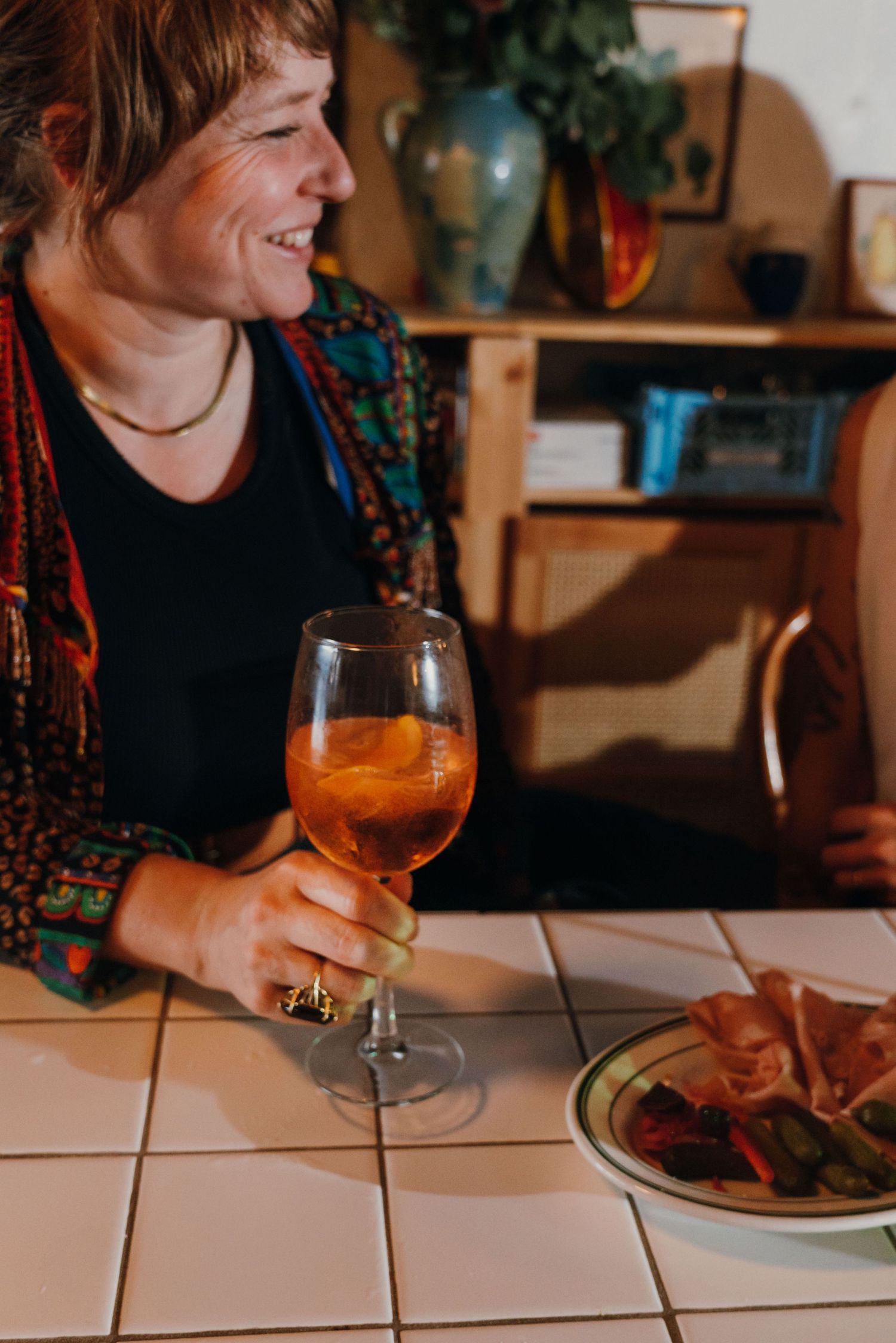 Smiling woman holding an Aperol spritz cocktail beside a charcuterie plate at a tiled bar counter.