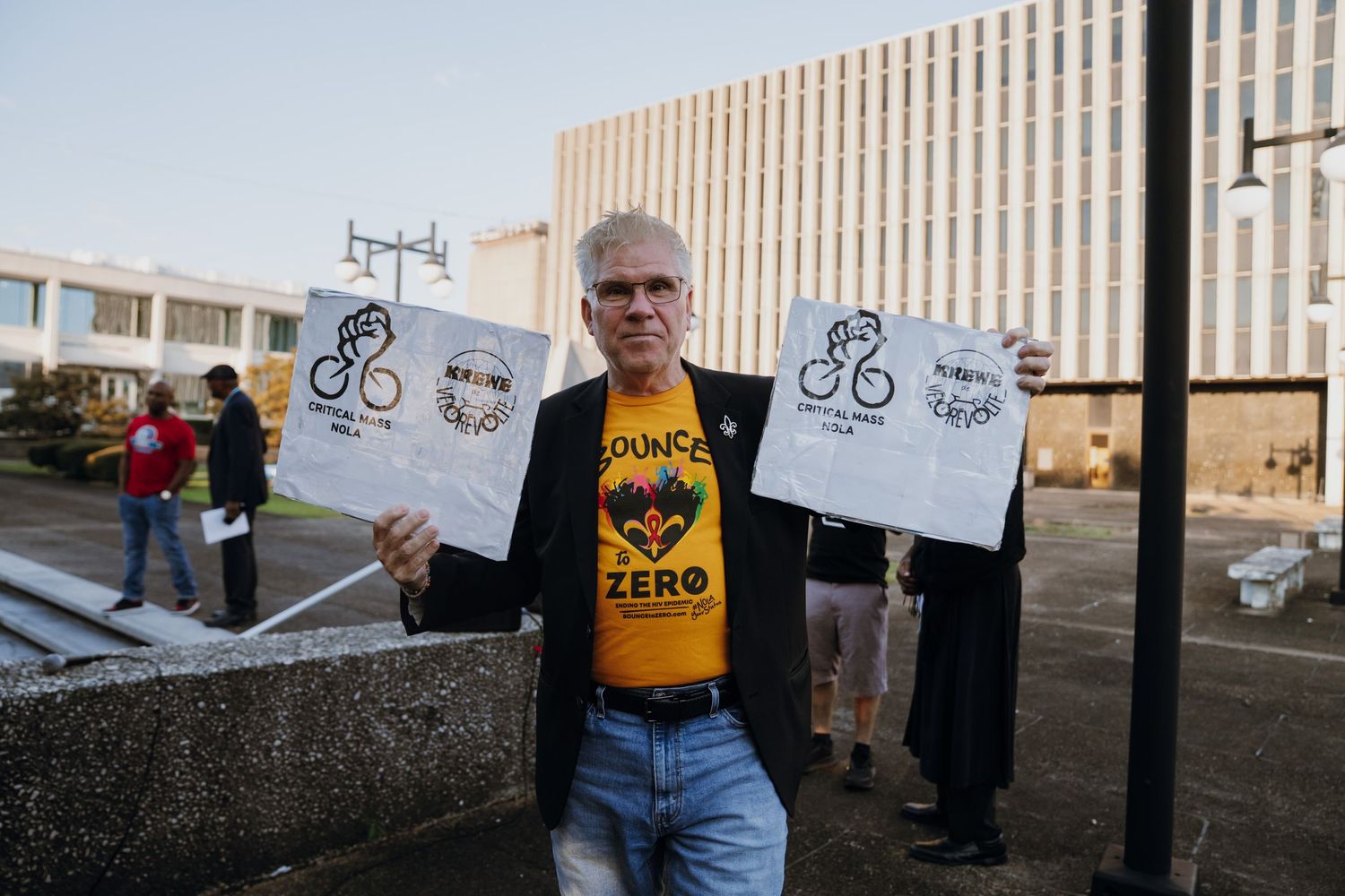 Man in yellow 'Bounce Zero' shirt holding protest signs outside government building.