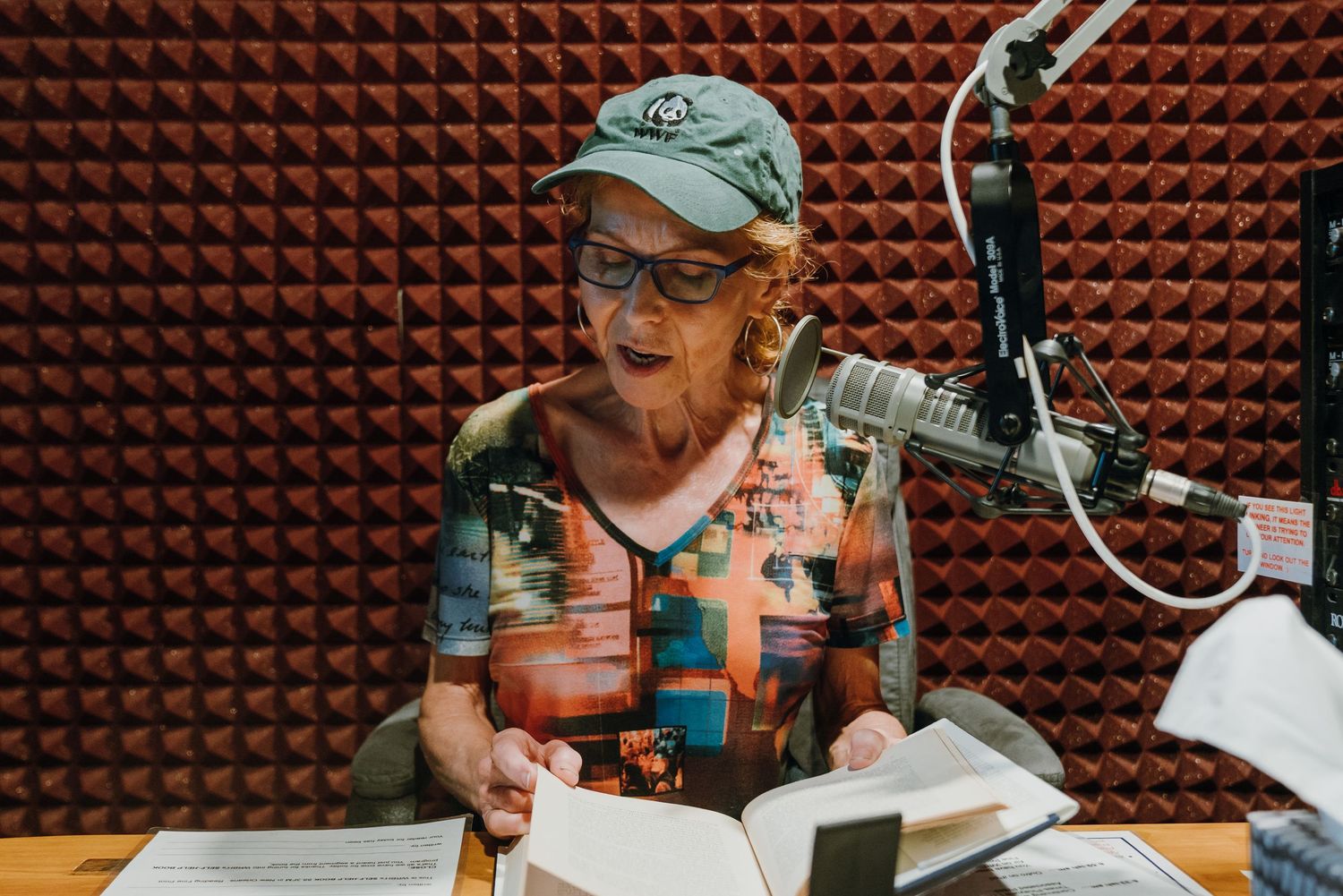 Woman reading a book aloud into a microphone in a red acoustic foam-lined recording studio booth.