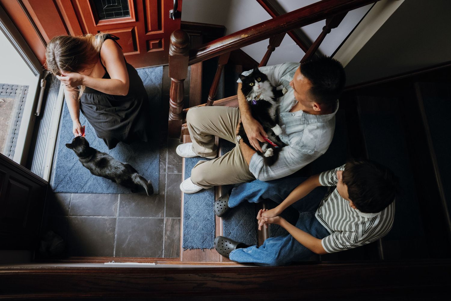Overhead view of a family sitting on stairs interacting with a black and white cat indoors.