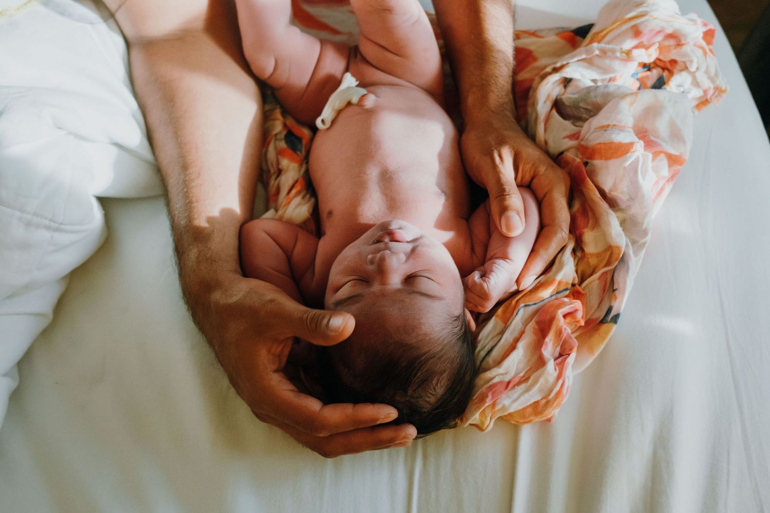 Newborn baby lying on white bed being gently held by parents hands wrapped in floral swaddle. New Orleans, Louisiana, Mississippi