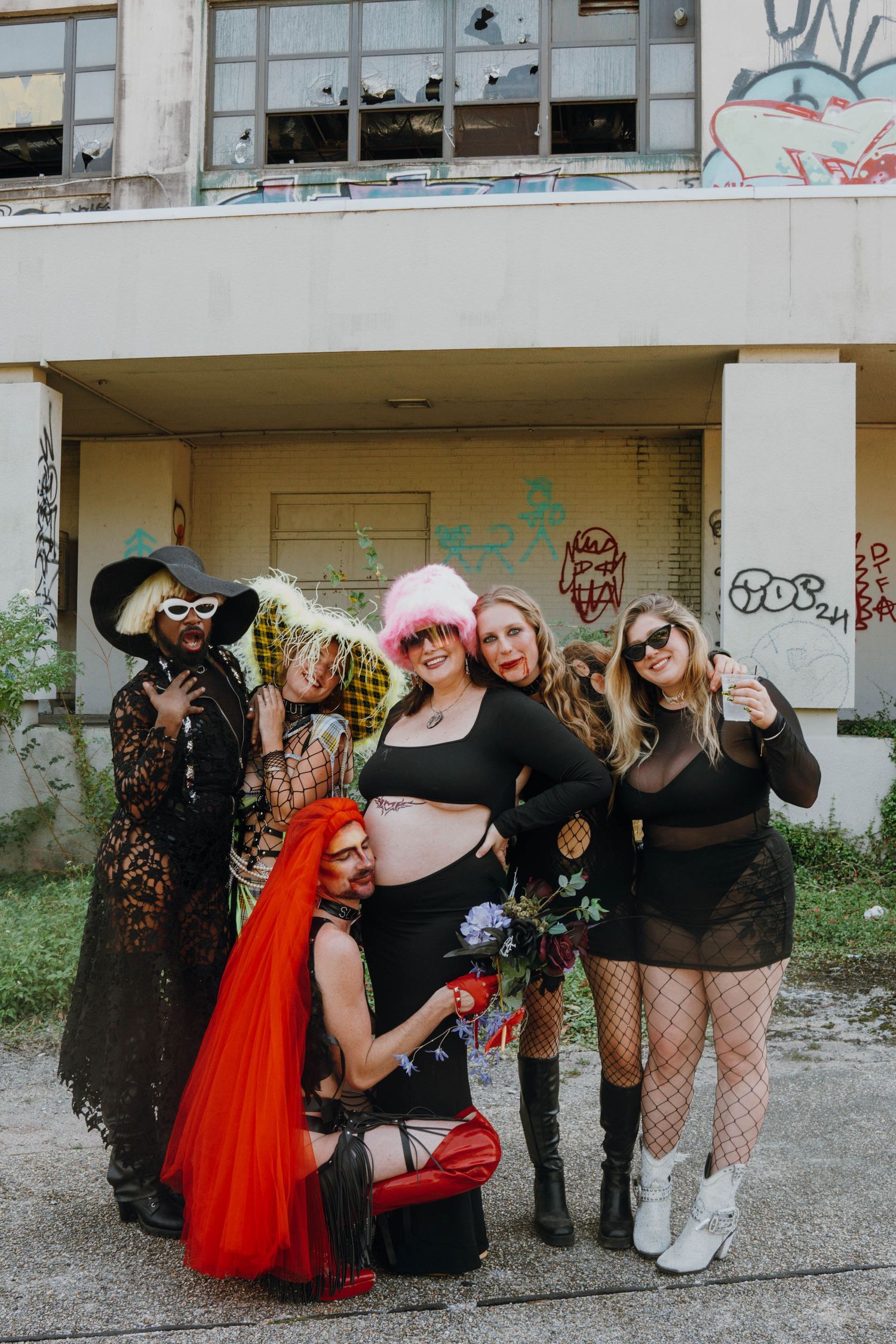 Group of six people in creative costumes posing before a graffiti-covered abandoned building.