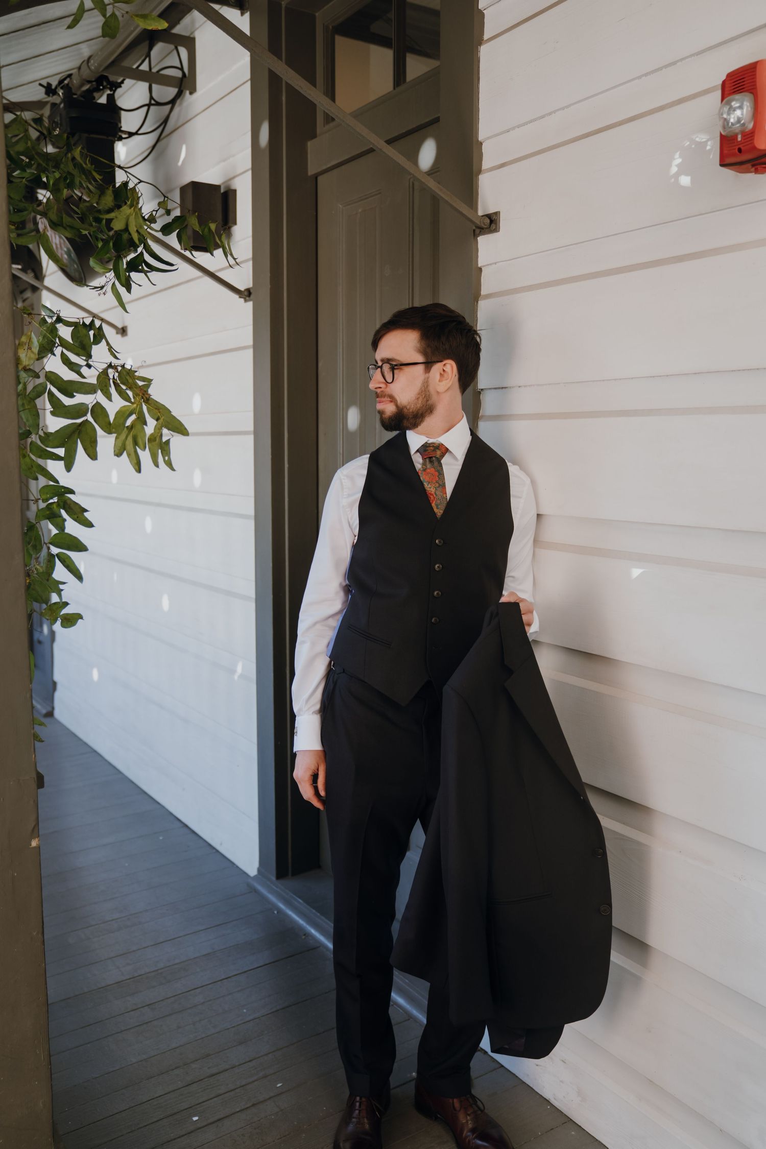 Groom in dark vest and tie holding suit jacket on white building porch before wedding.