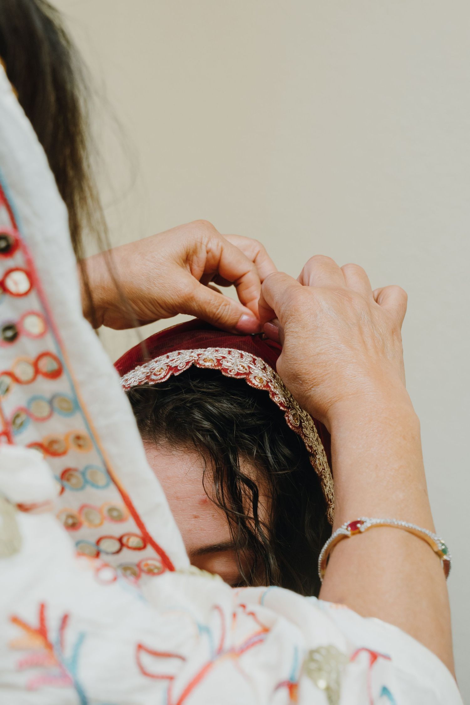 Hands adjusting a decorative red and gold headpiece on a bride's dark hair during Indian wedding preparations.