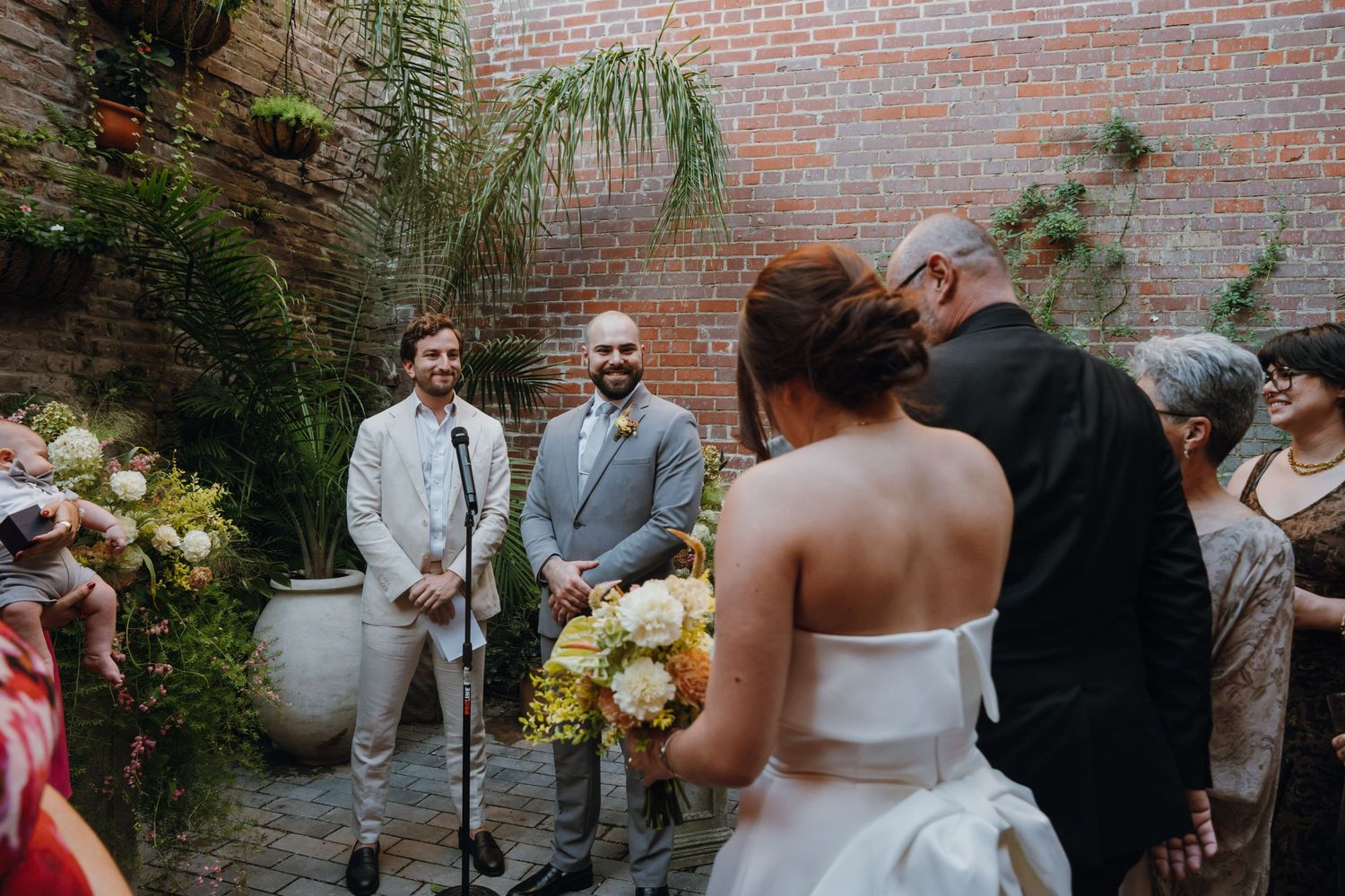 Groom in gray suit smiles as bride approaches outdoor wedding ceremony at rustic brick courtyard venue. New Orleans, Louisiana, Mississippi
