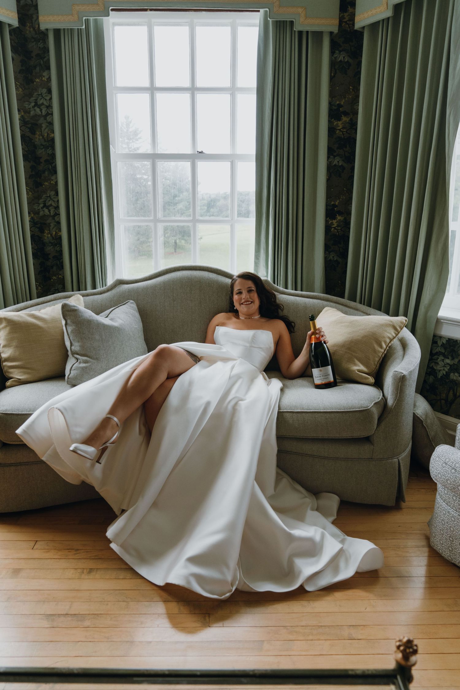 Smiling bride in strapless white wedding gown relaxing on a sofa holding a champagne bottle. New Orleans, Louisiana, Mississippi