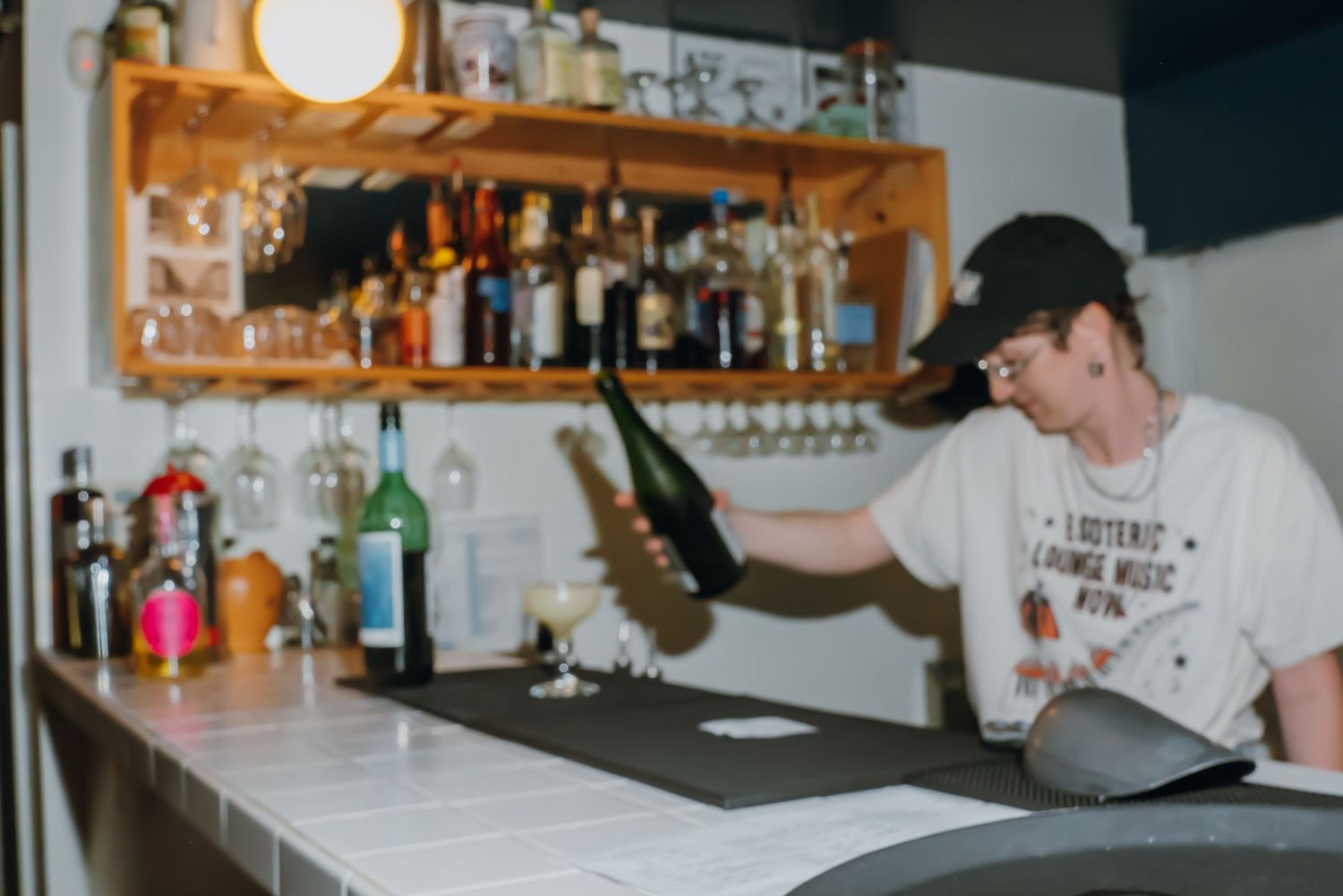 Bartender holding a wine bottle behind a tiled bar counter with liquor shelf in background.