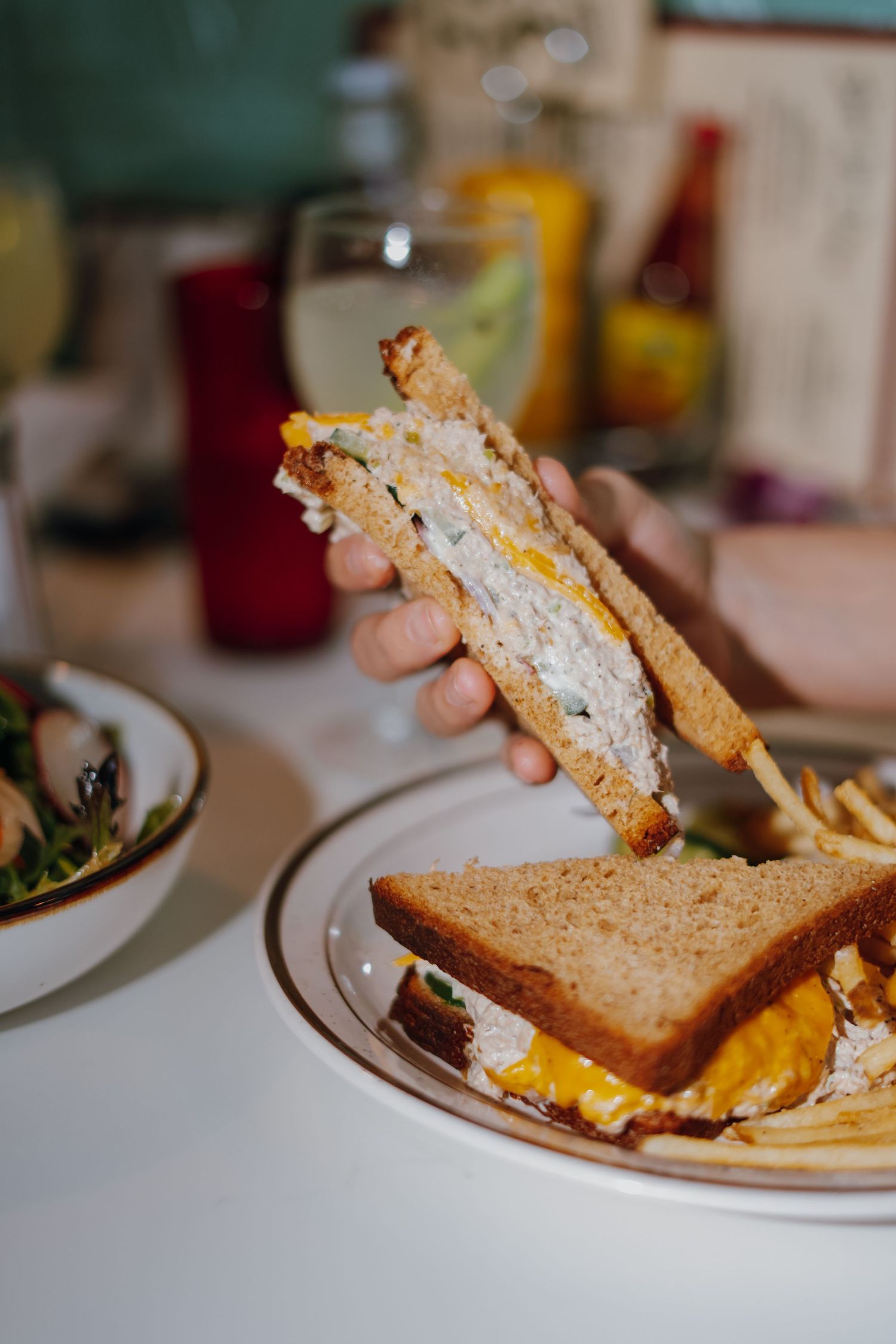 Hand holding half of a tuna salad sandwich on wheat bread over plate with fries.