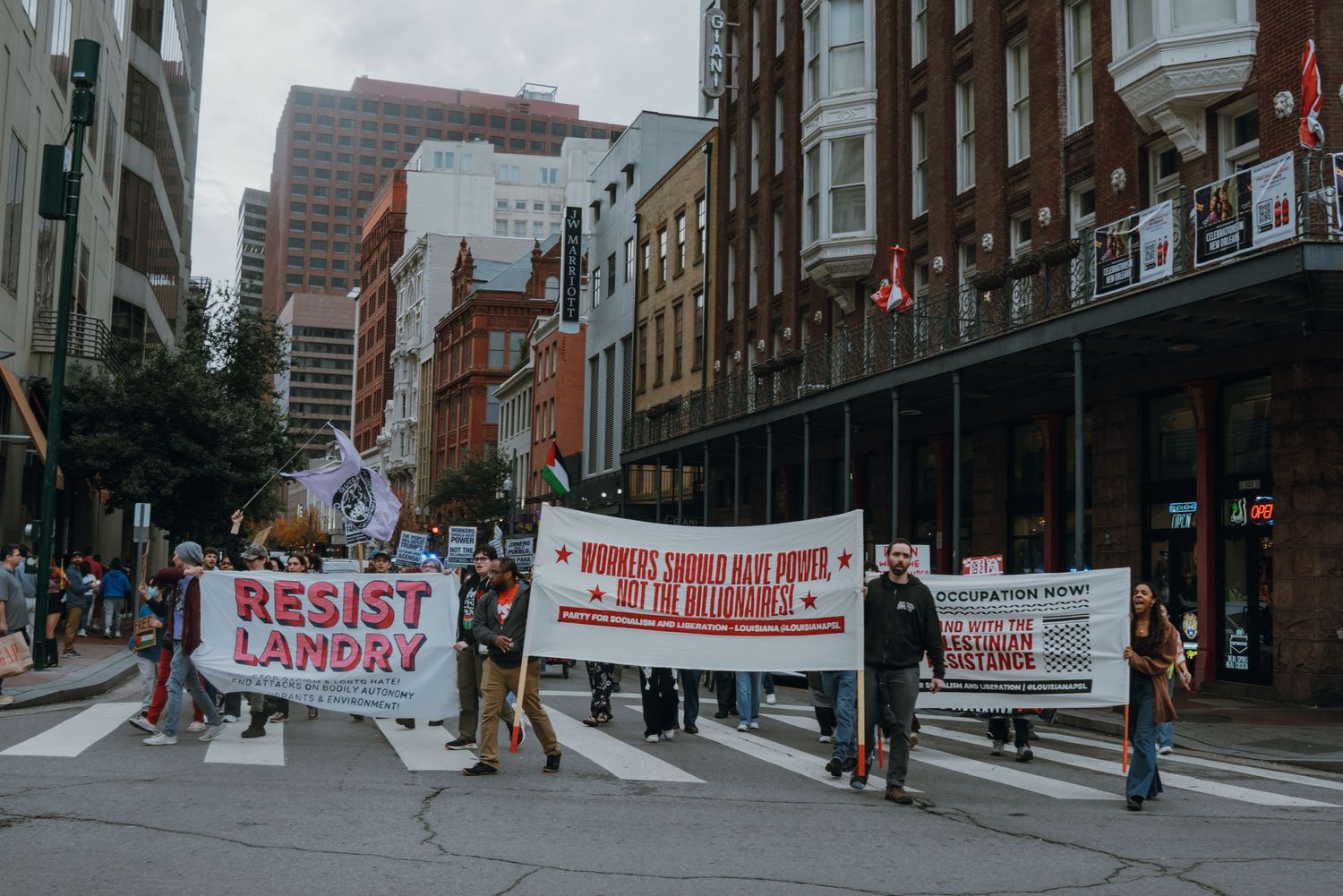 Protesters carry a red Resist Landry banner while marching through a historic urban street.