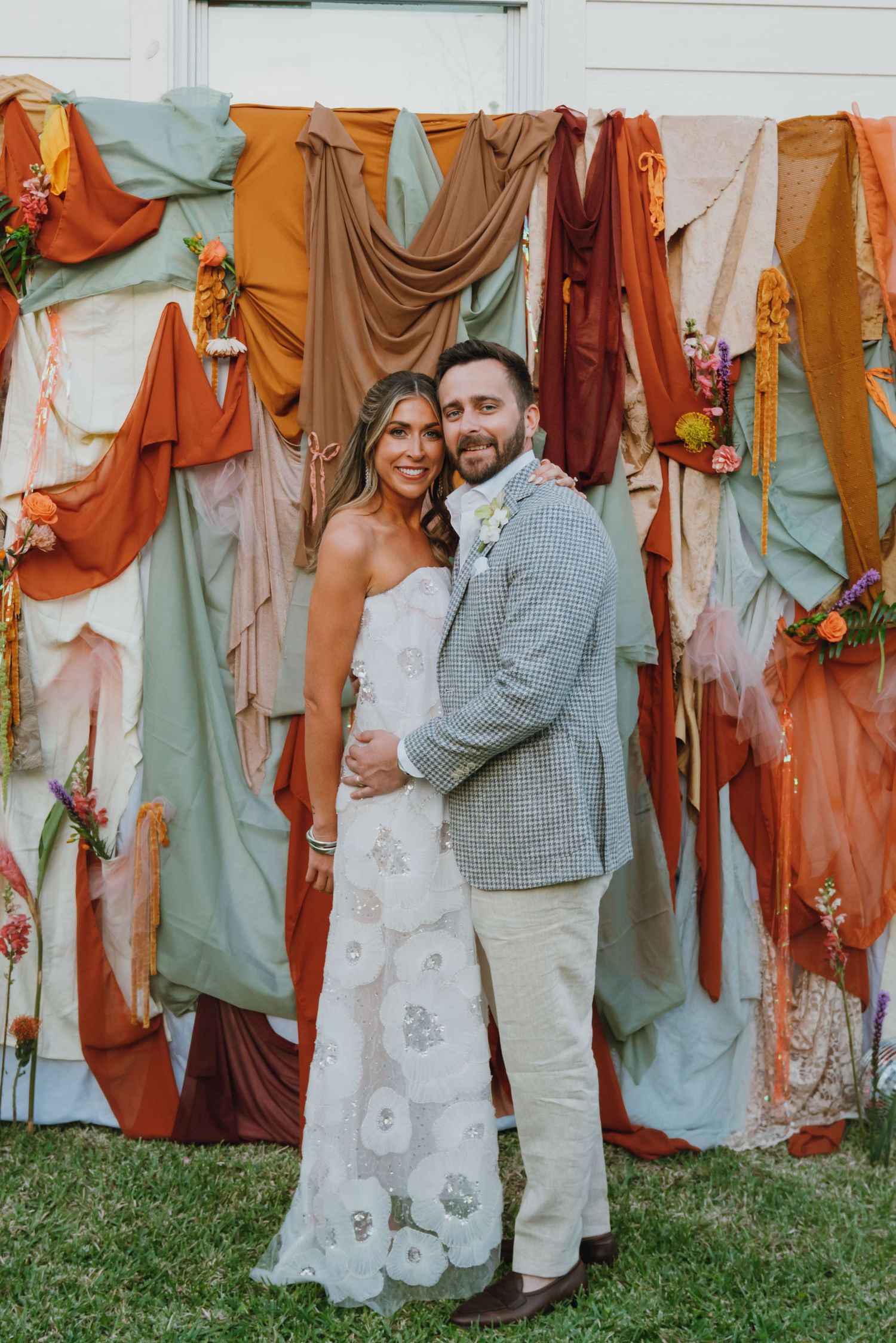 Bride and groom pose before a colorful draped fabric backdrop with floral accents at outdoor wedding reception.