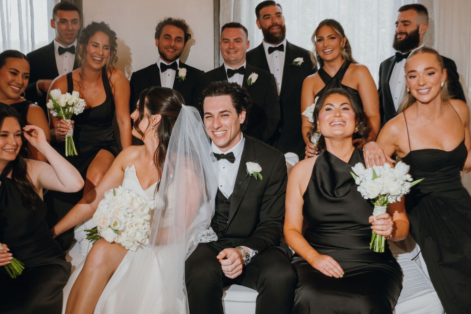 Wedding party in black attire laughs together with bride in white gown and groom in tuxedo.