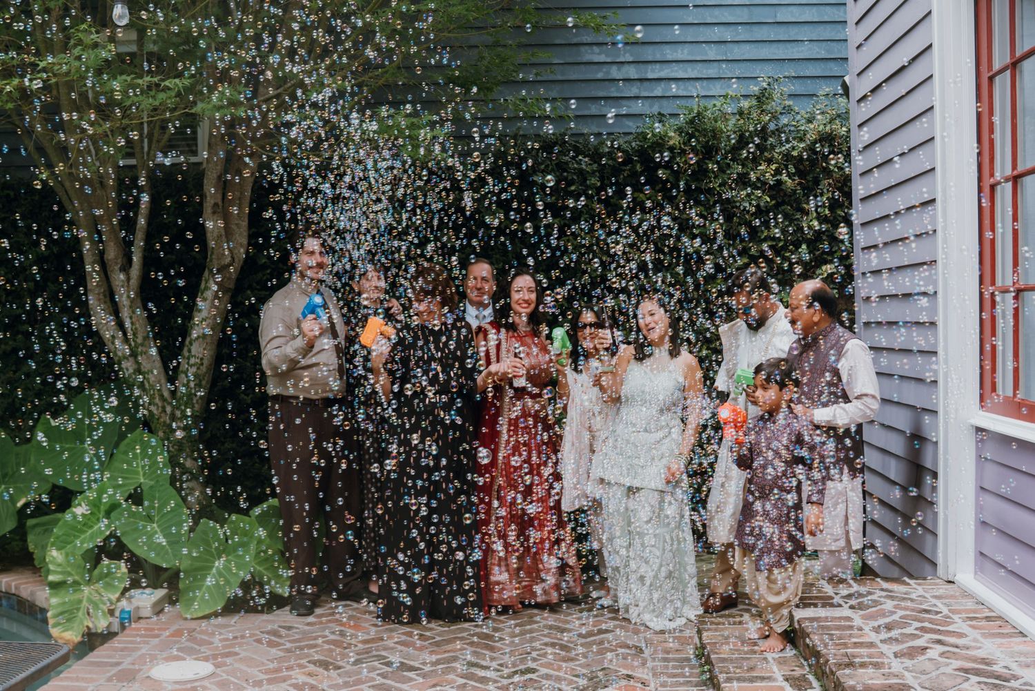 Wedding party celebrating with bubble guns outside a charming blue house on brick pathway.