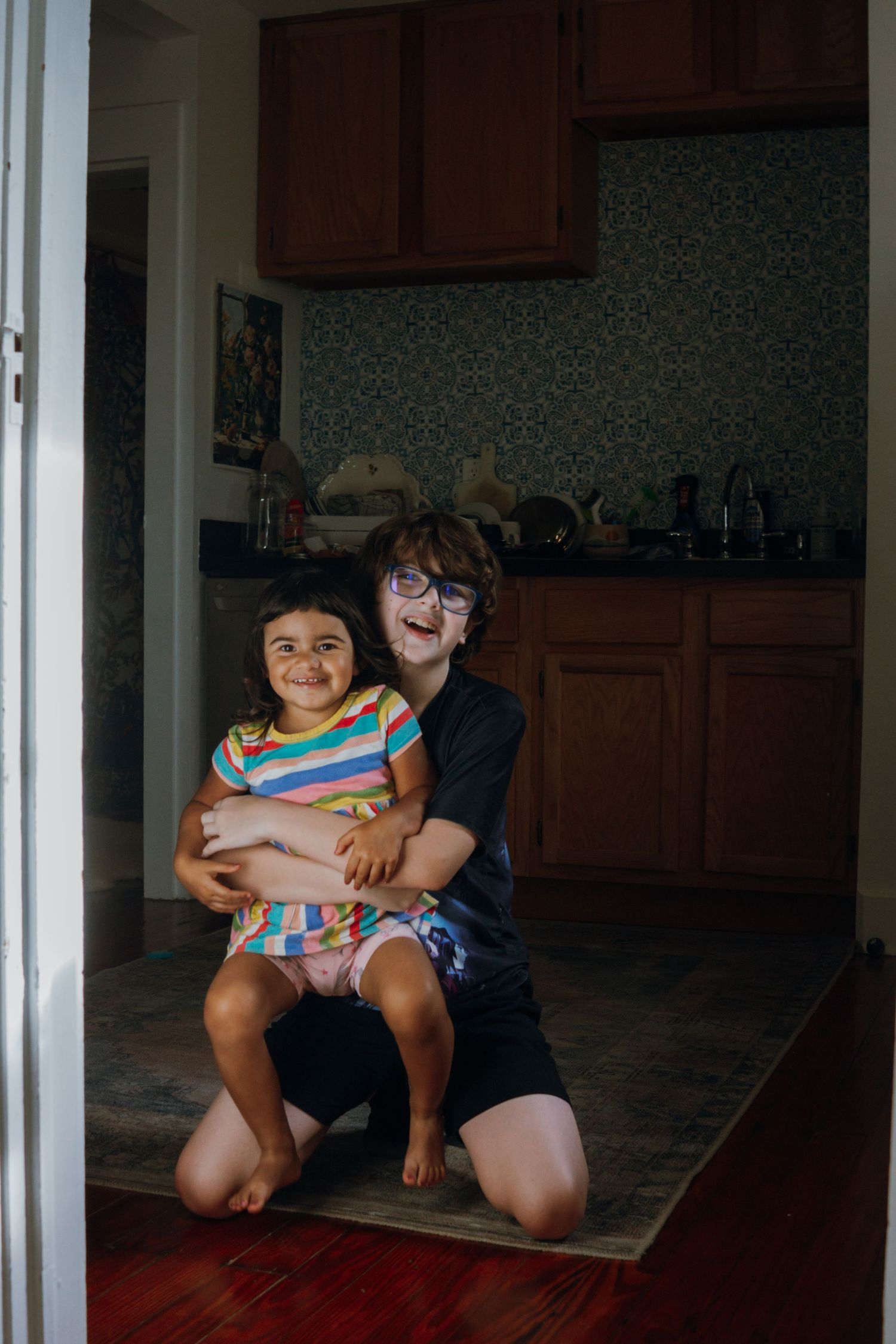 Two smiling children hugging on kitchen floor, older child kneeling behind younger in colorful striped shirt.
