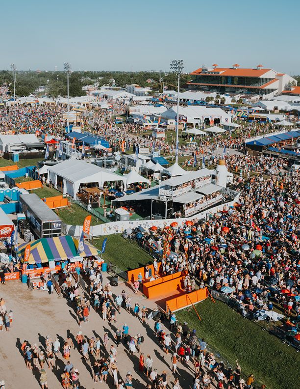 Aerial view of a large outdoor festival with massive crowds, colorful tents, and orange-accented stages on a sunny day.